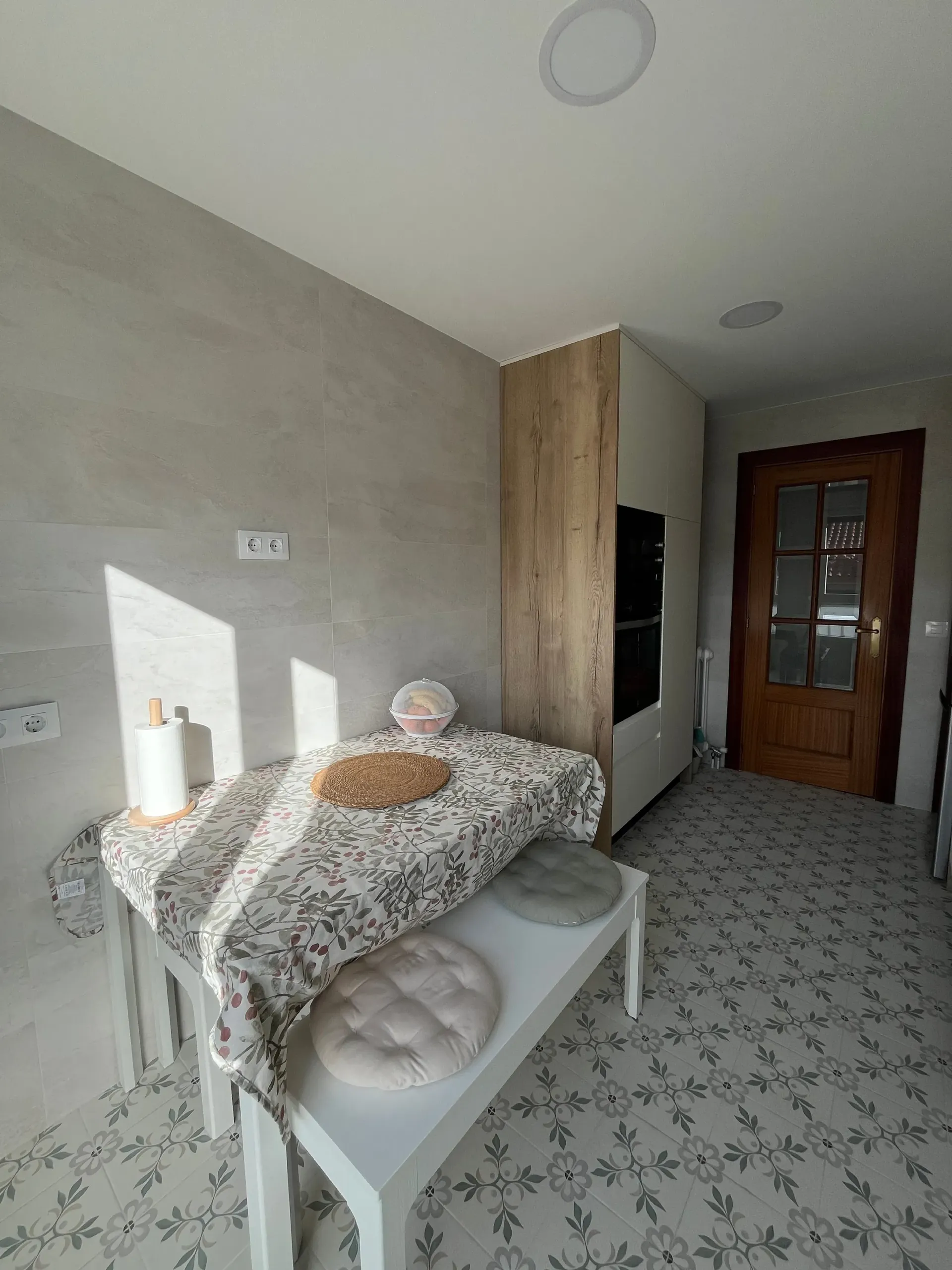 Una mesa blanca con tela estampada y cojines se encuentra en una habitación con una pared gris claro, un mueble revestido de madera y suelo de baldosas.