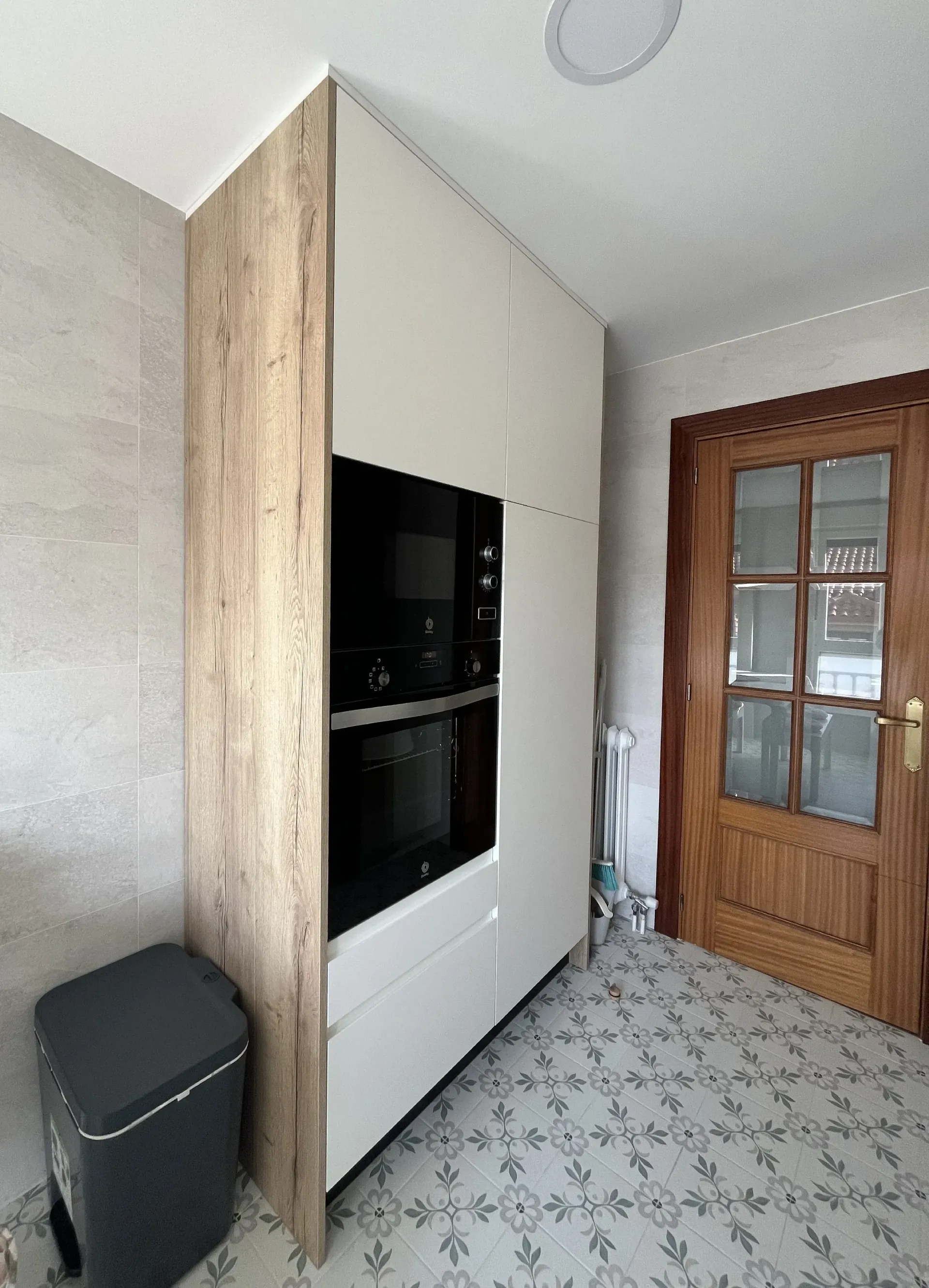 Mueble de cocina empotrado con armarios blancos, panel lateral con acabado de madera y horno y microondas apilados en color negro.
