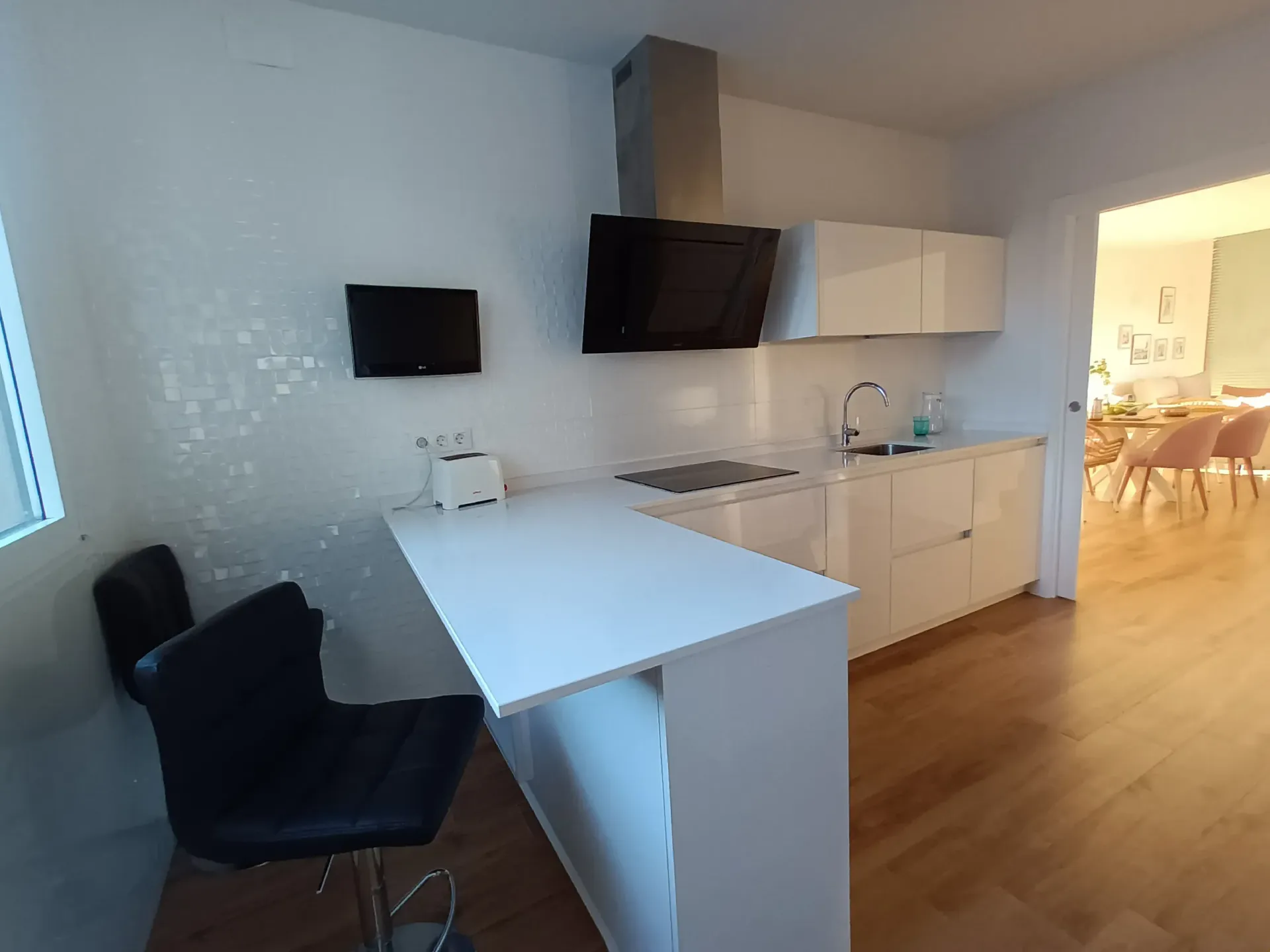 Una cocina moderna con armarios blancos, campana extractora negra, barra de desayuno blanca con silla negra y suelo con aspecto de madera.