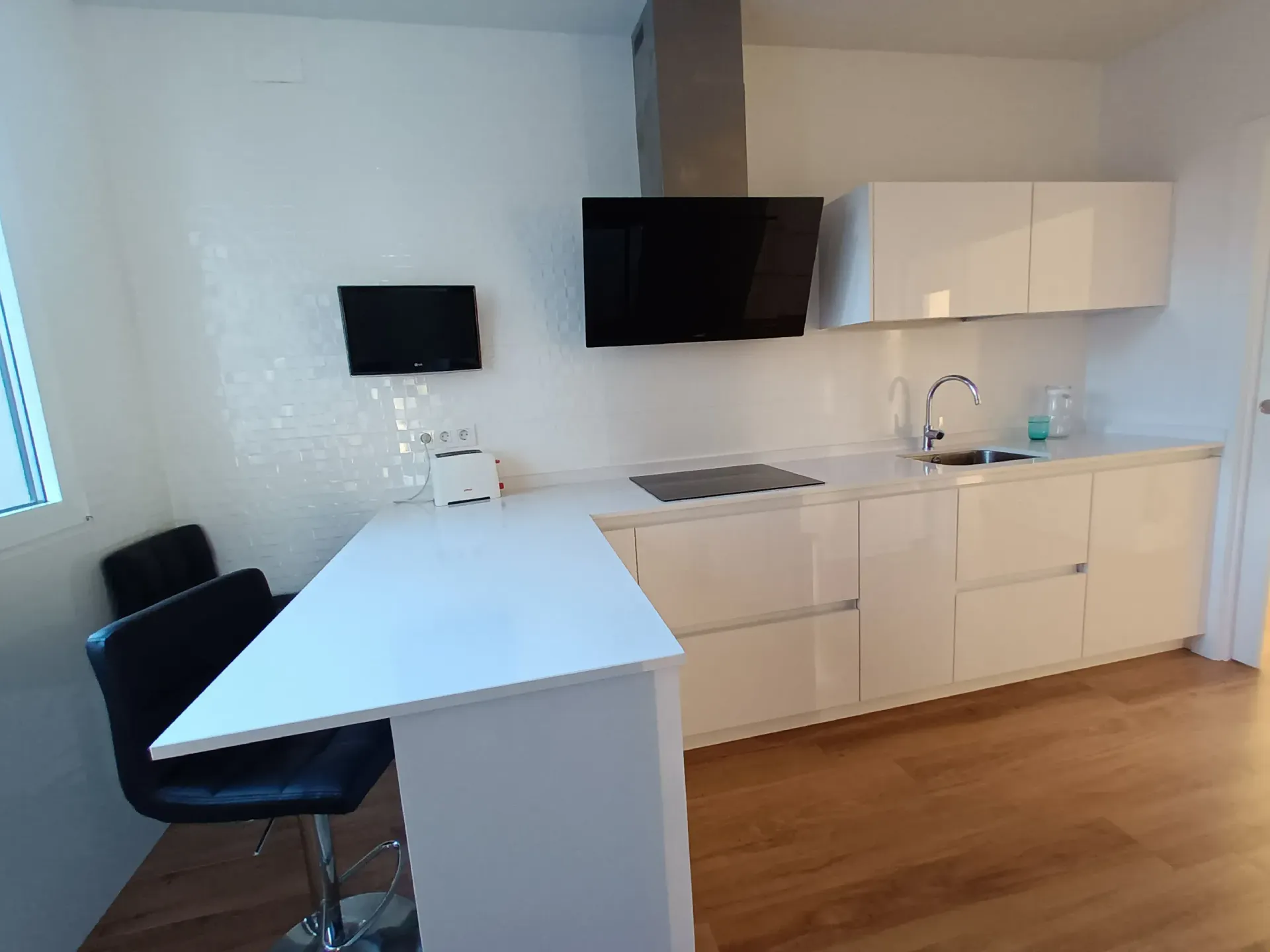 Una cocina moderna con armarios blancos, campana extractora negra, barra de desayuno en la isla y dos taburetes negros sobre suelo de madera.