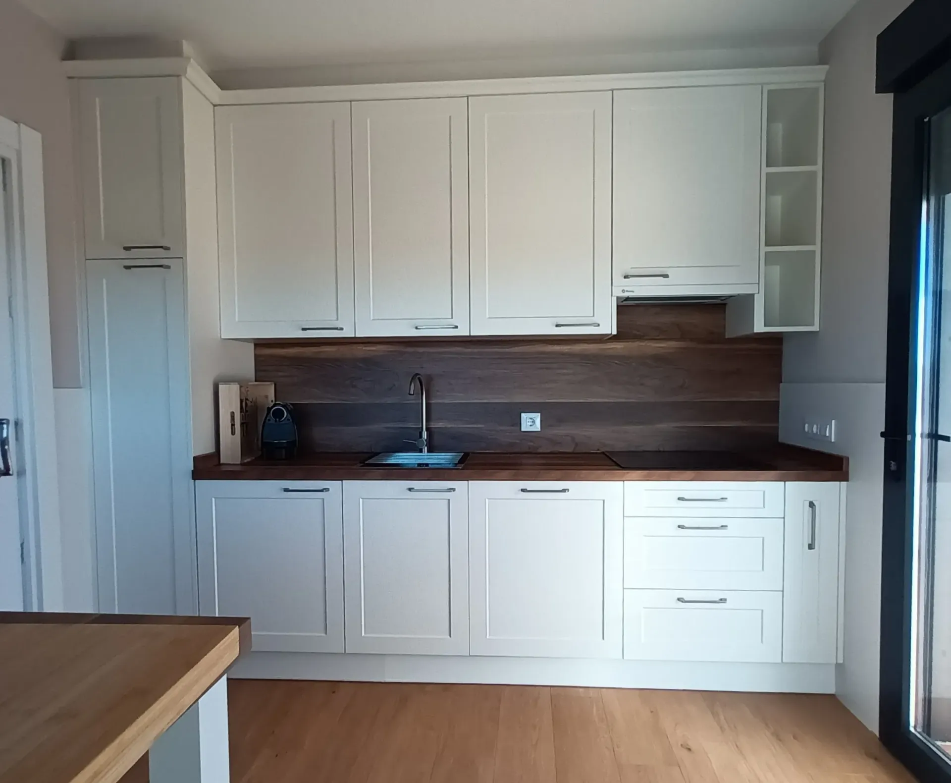 Cocina moderna con armarios blancos, encimeras y salpicadero de madera, y suelo de madera clara en un espacio luminoso y compacto.