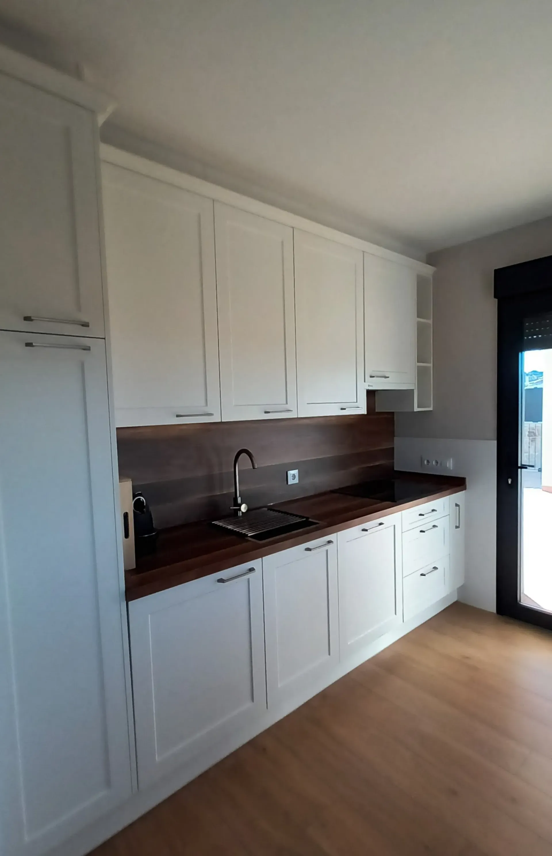 Una cocina moderna con armarios blancos, encimera de madera oscura, fregadero de acero inoxidable y suelo de madera clara.