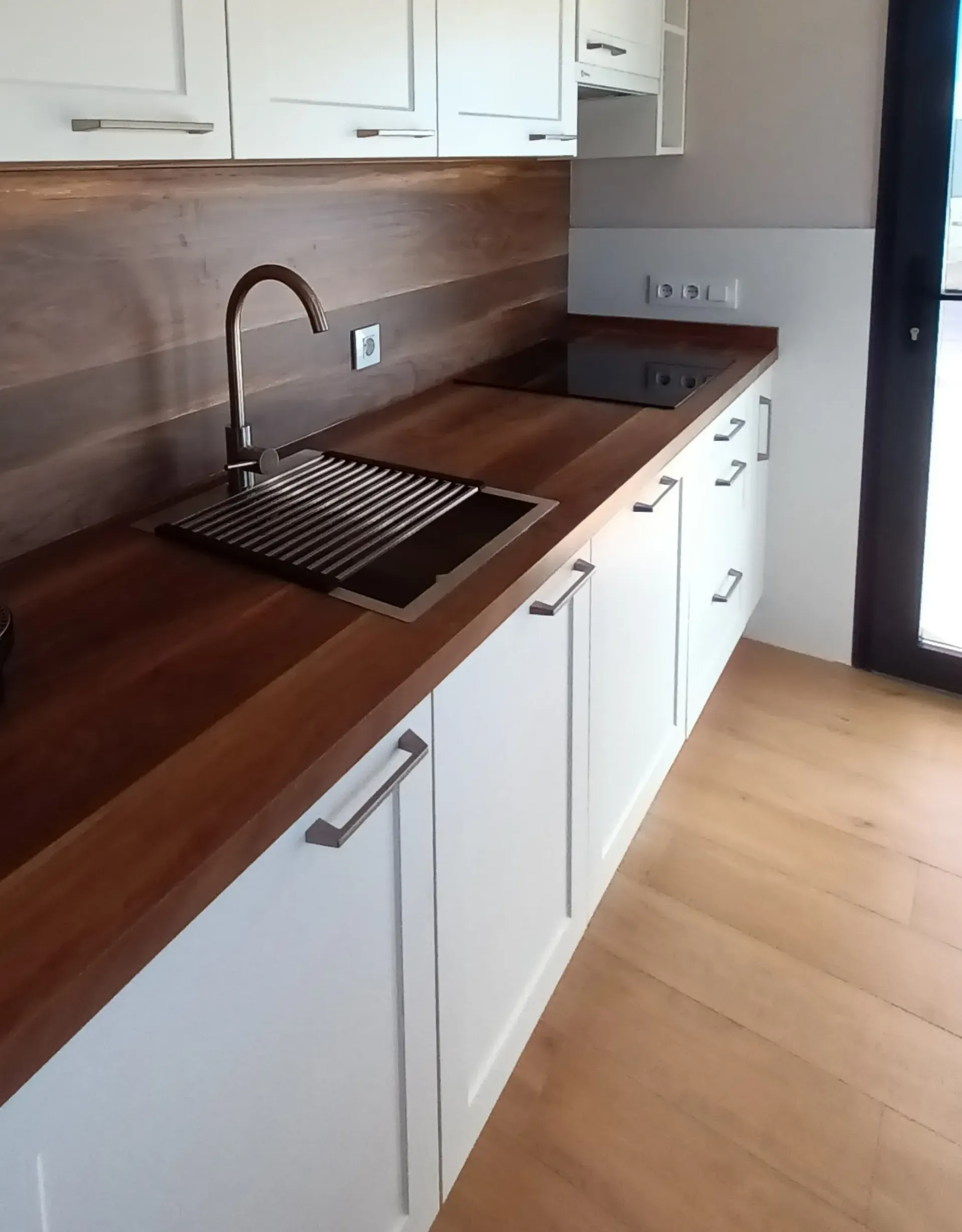 Una cocina moderna con armarios blancos, encimera de madera oscura, fregadero de acero inoxidable y placa de inducción negra.