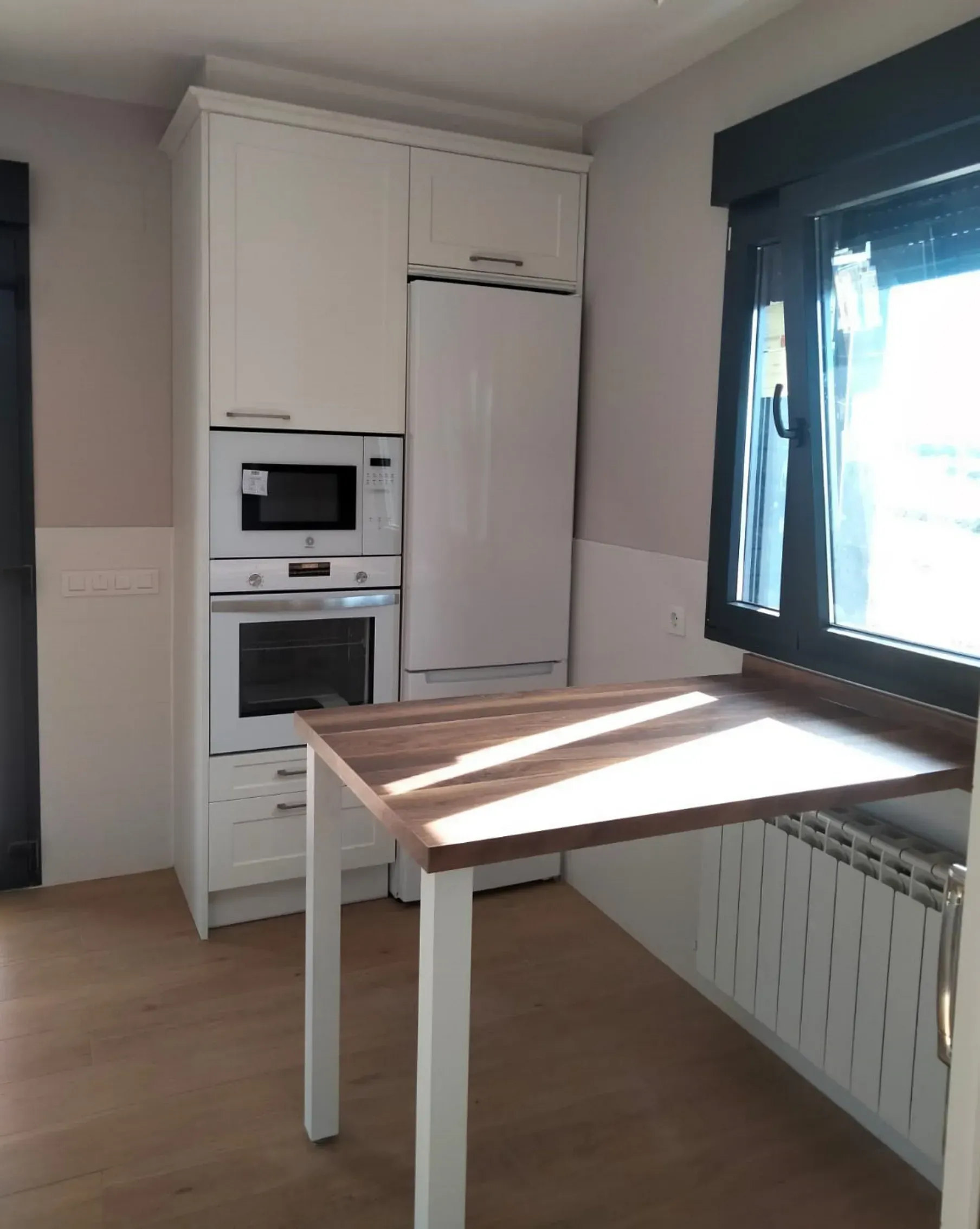 Un mueble de cocina blanco con microondas y horno integrados, junto a una barra de desayuno con encimera de madera, al lado de una ventana.