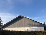 Haus mit grauem Giebeldach und weißen Wänden, teilweise hinter einer Hecke unter blauem Himmel versteckt