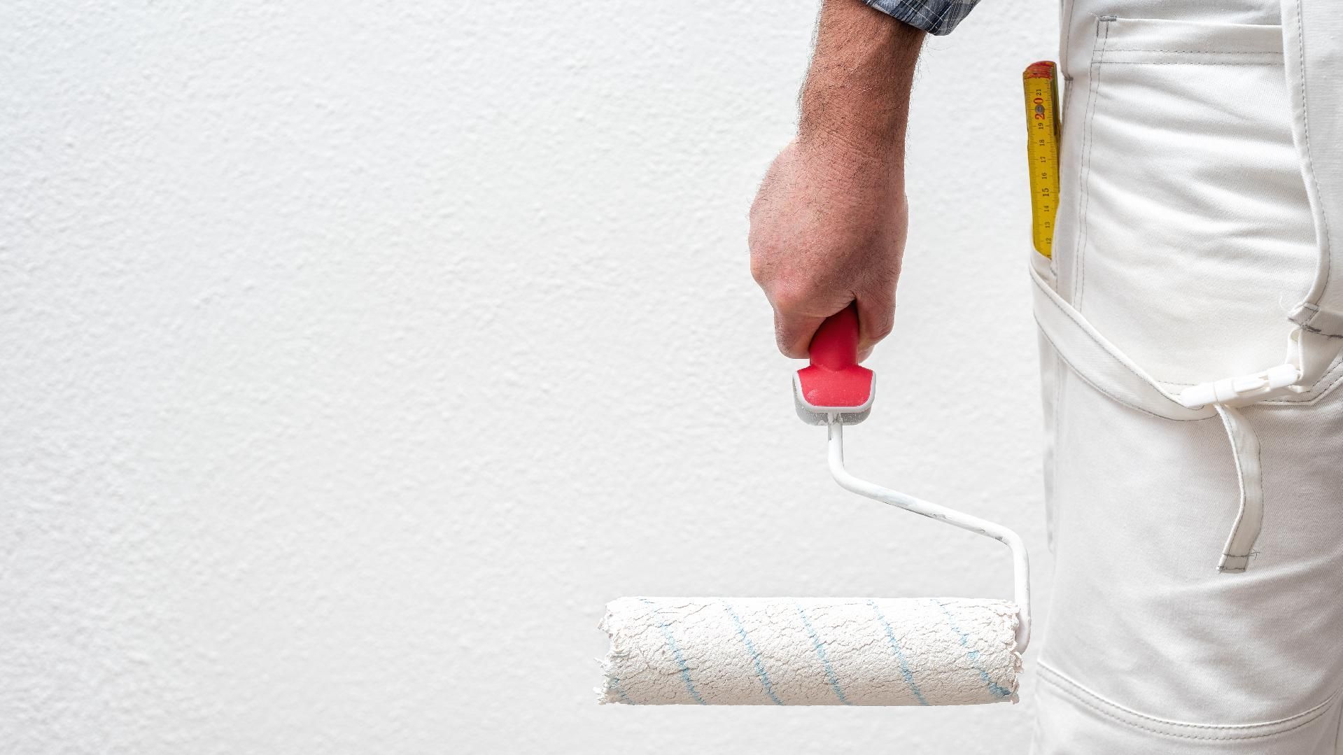 Eine Person in weißer Arbeitshose hält eine Farbrolle an eine schlichte weiße Wand.