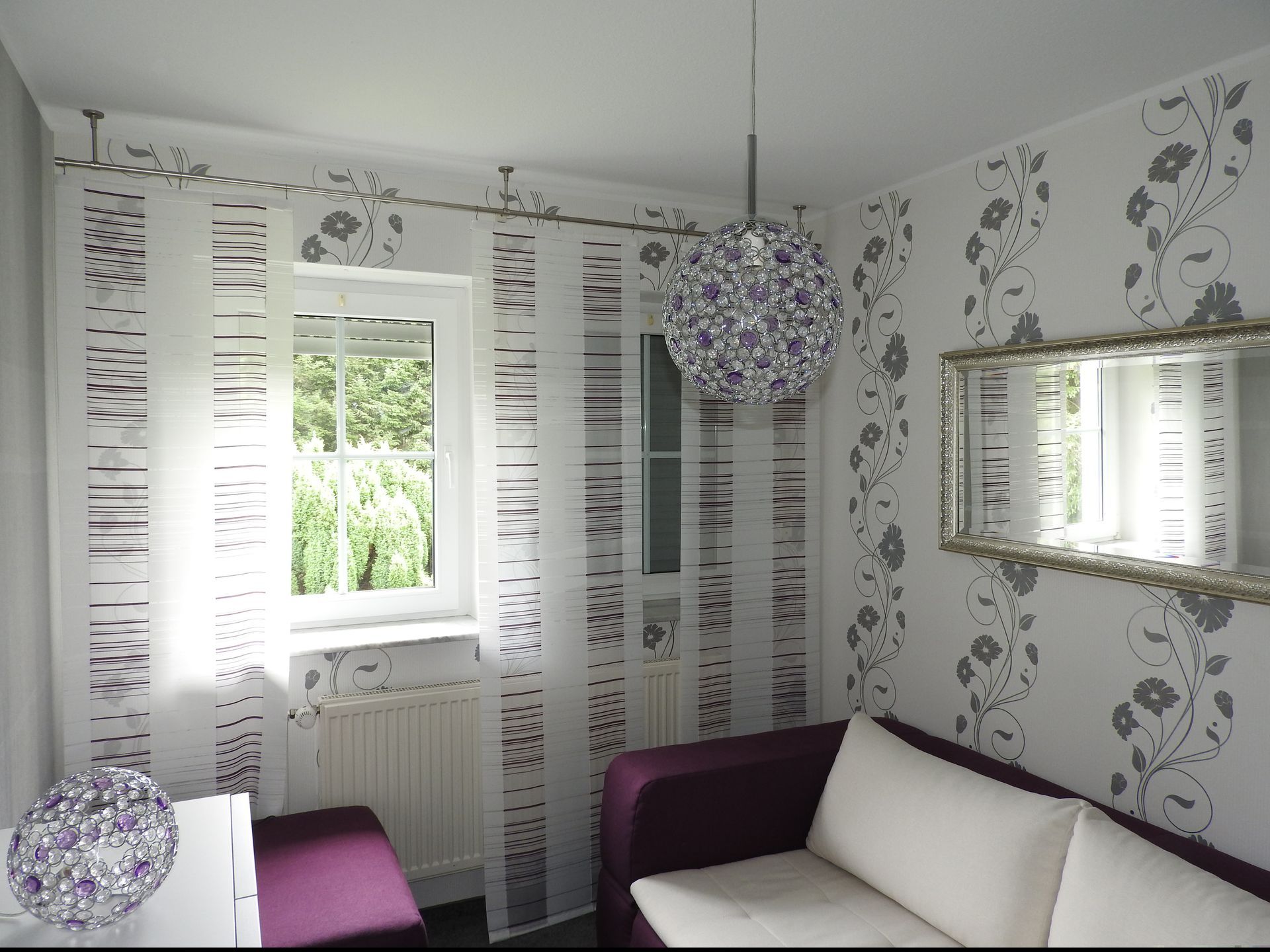 Ein Eckzimmer mit weißer Tapete mit Blumenmuster, einem weißen Sofa mit violetten Akzenten und einer Pendelleuchte.