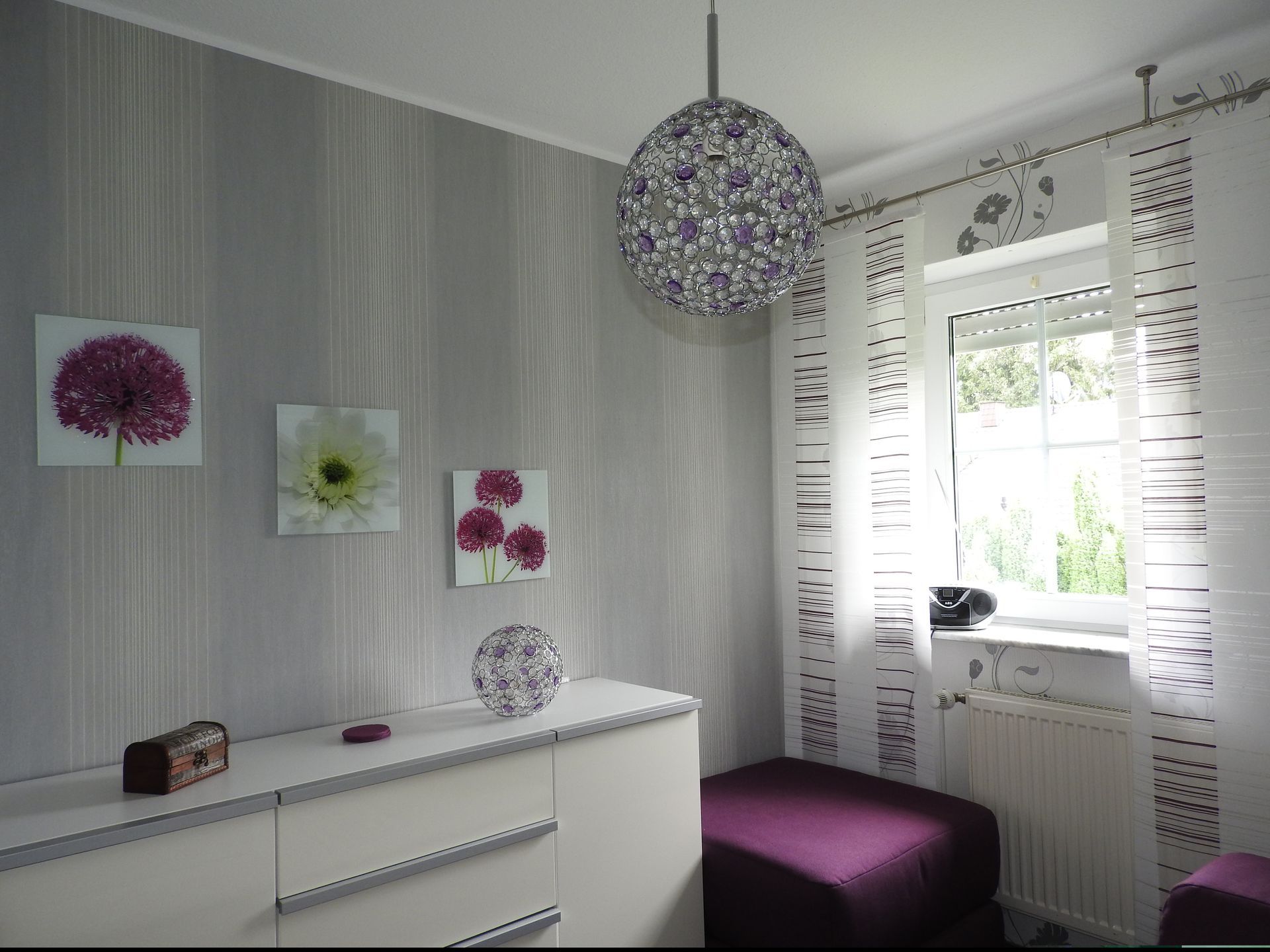 Modernes Zimmer mit grauer Mustertapete, floralen Leinwanddrucken, Kugel-Pendelleuchte und lilafarbenem Ottoman.