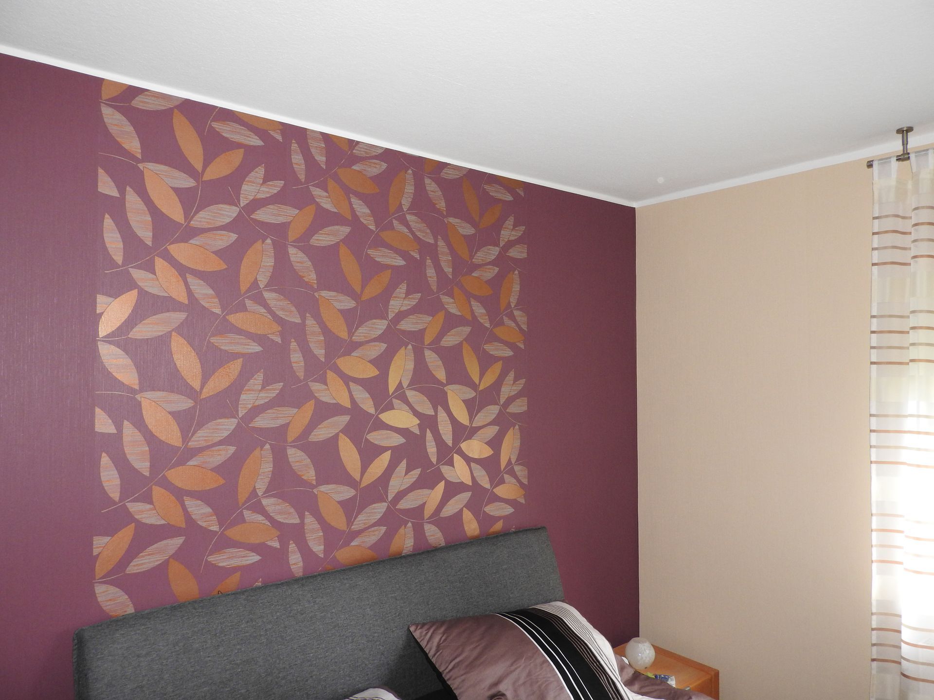 Pflaumenfarbene Wand mit goldenem Blättermuster und stilvollem grauen Kopfteil. Ein eleganter Akzent.