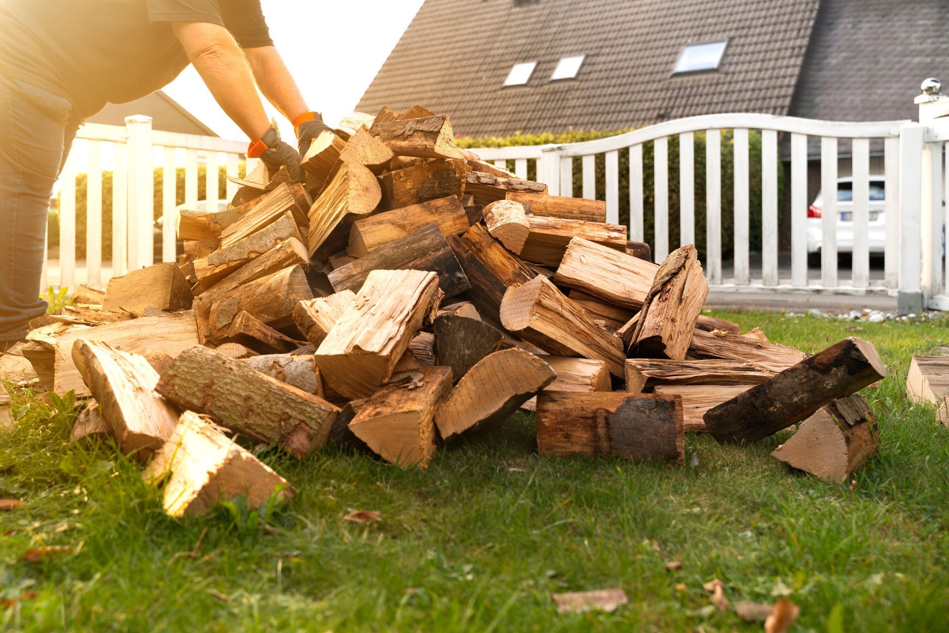 Une personne empile du bois de chauffage sur une pelouse près d'une clôture blanche.