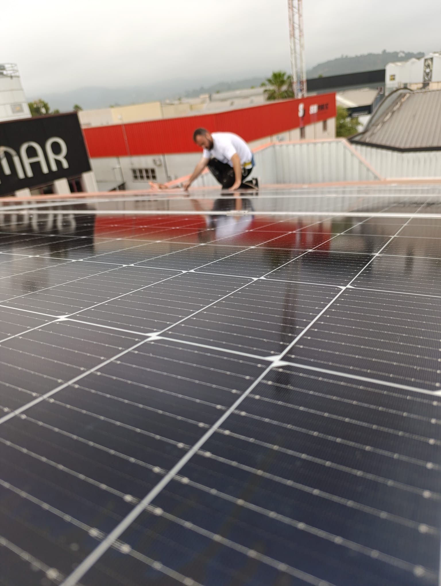 Persona limpiando paneles solares en un tejado. Edificio rojo brillante al fondo. Día nublado.