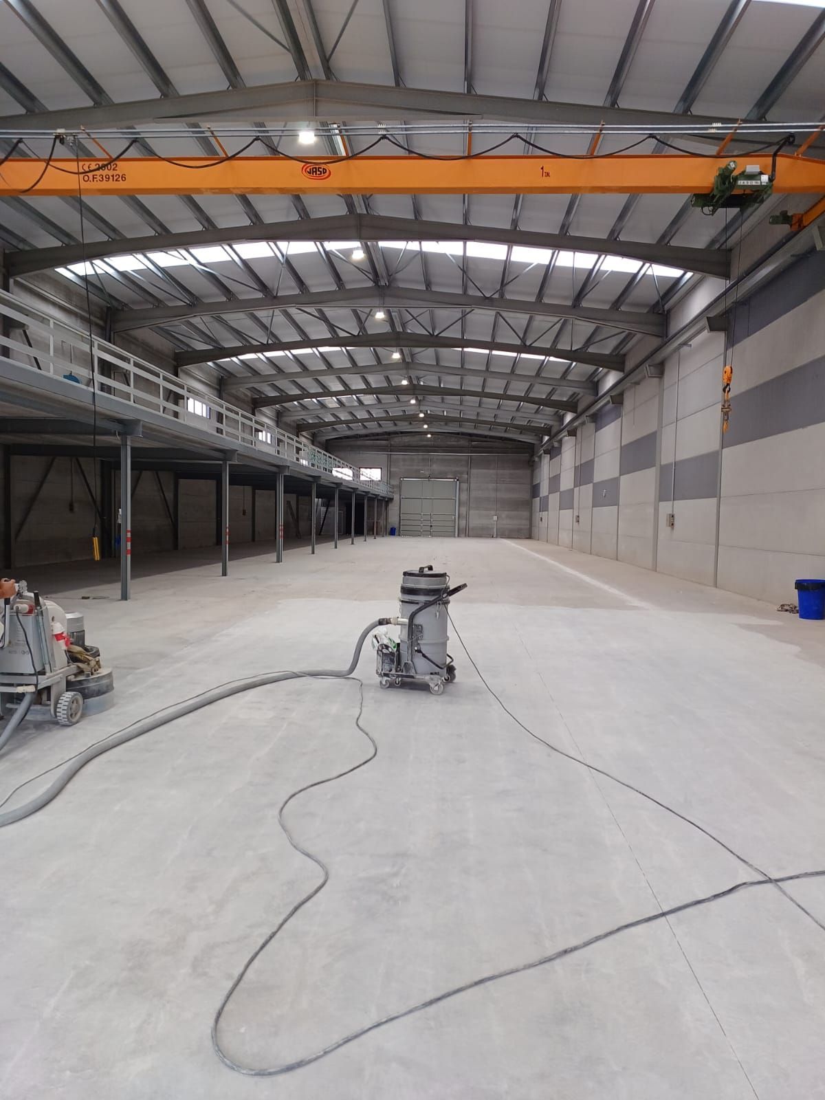 Gran nave industrial vacía con suelo de hormigón. Una máquina de preparación de superficies se encuentra en el centro.