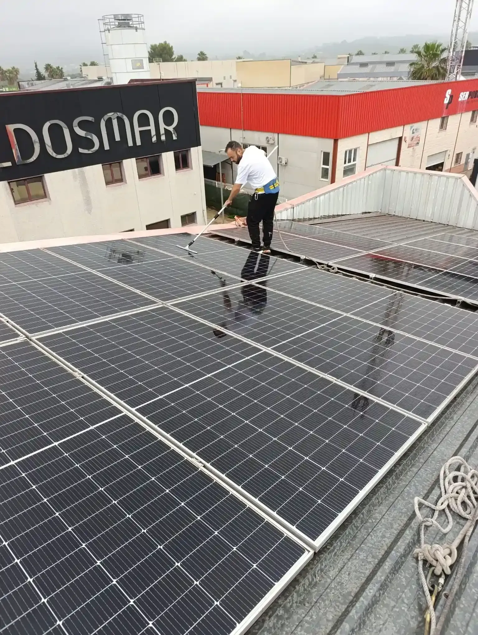Hombre limpiando paneles solares en un tejado, con arnés de seguridad. Día nublado, entorno industrial.