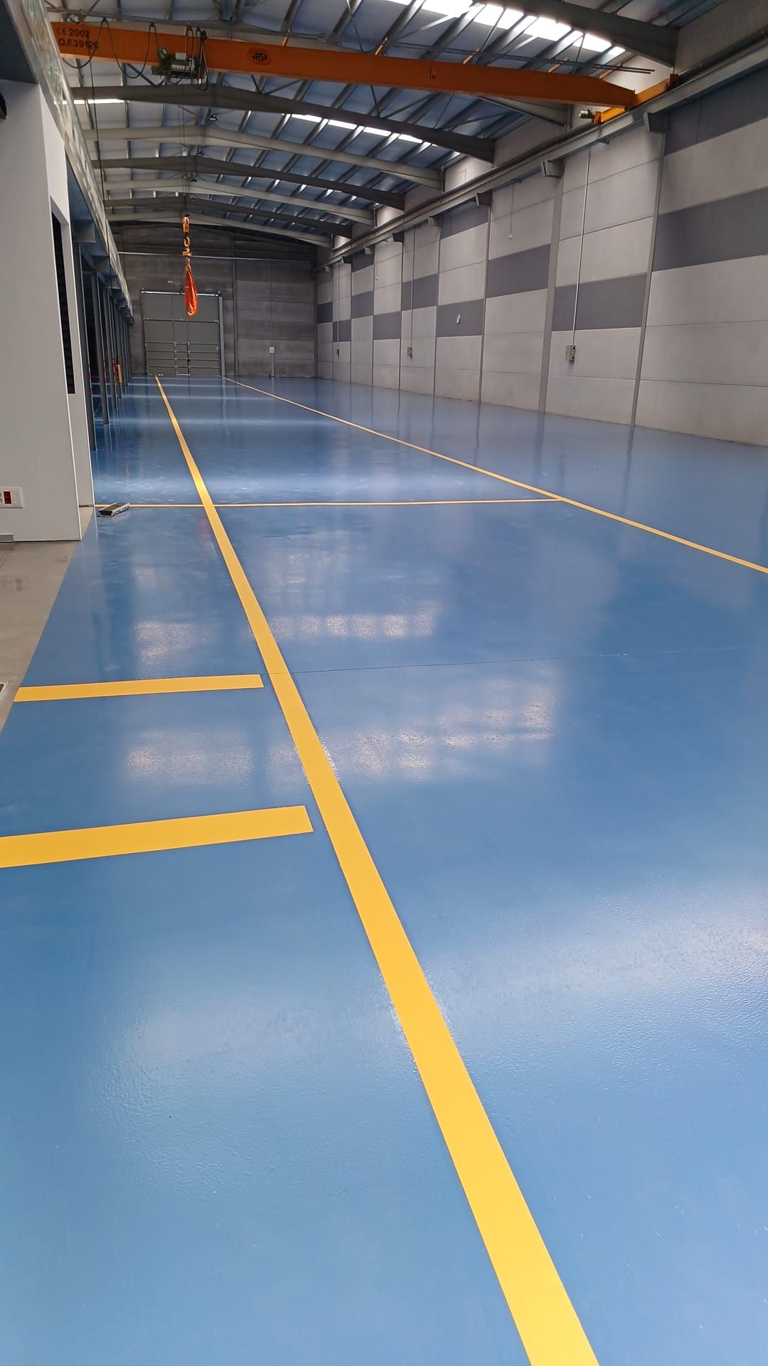 Piso industrial azul con líneas amarillas, una grúa y paredes grises.