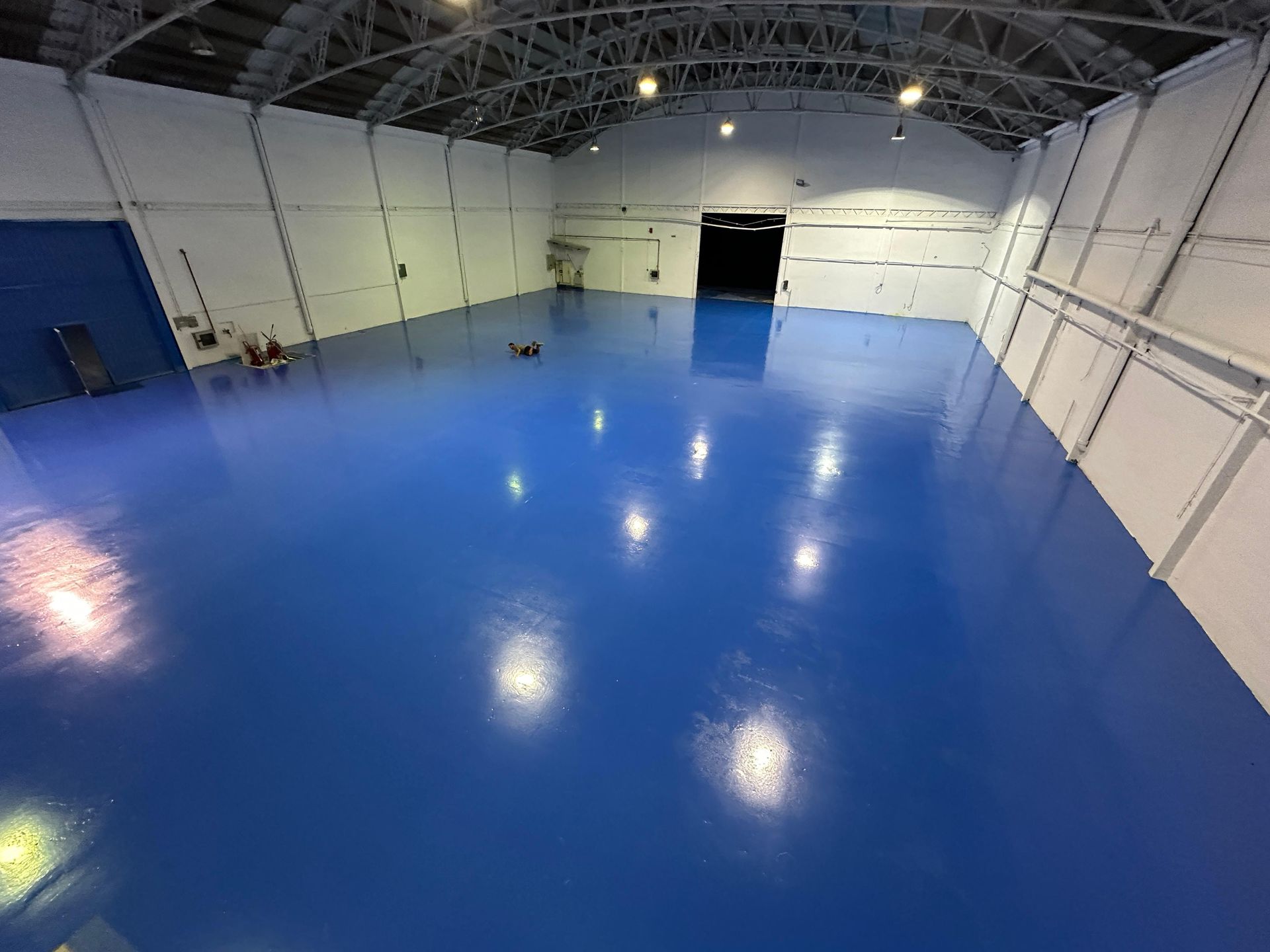 El interior de un almacén grande y vacío. Un suelo azul brillante refleja las luces del techo. Paredes blancas.
