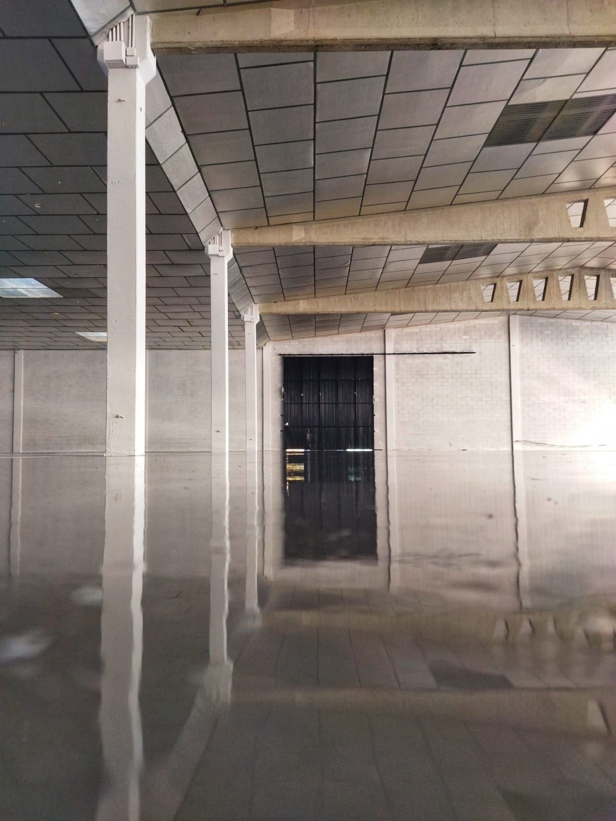 Vista interior de un edificio inundado. El agua refleja las tejas del techo y las vigas blancas. Al fondo hay una puerta metálica negra.