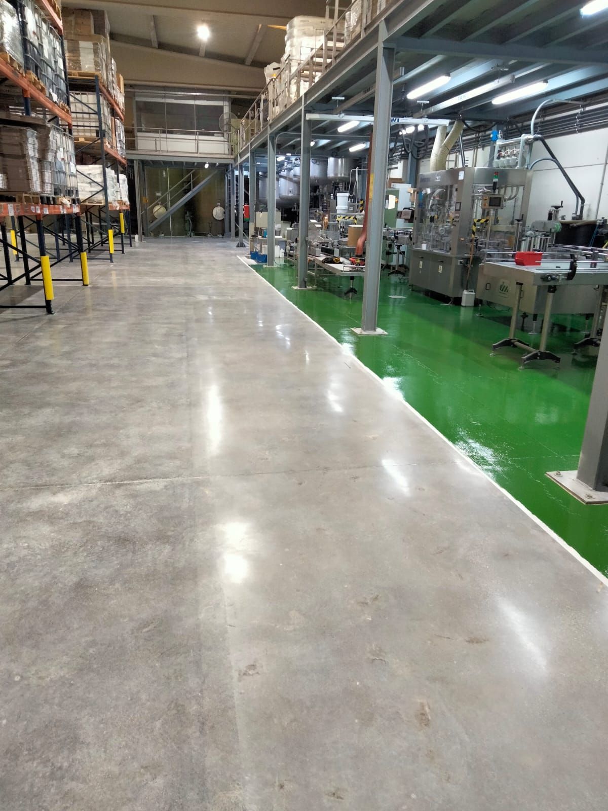 Interior de almacén, piso de concreto, línea de epoxi verde, maquinaria, almacenamiento elevado, iluminación brillante.