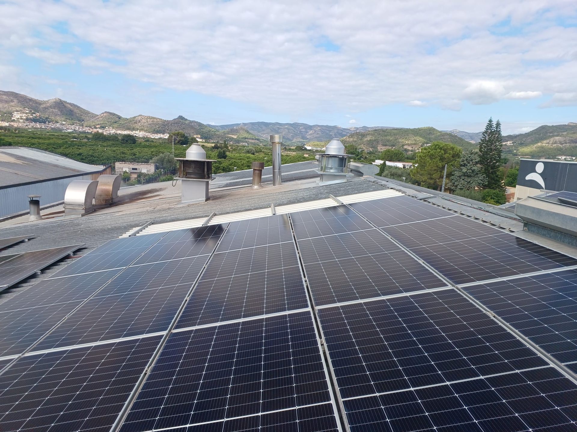 Paneles solares en un techo con tubos de escape, montañas y árboles al fondo bajo un cielo azul.
