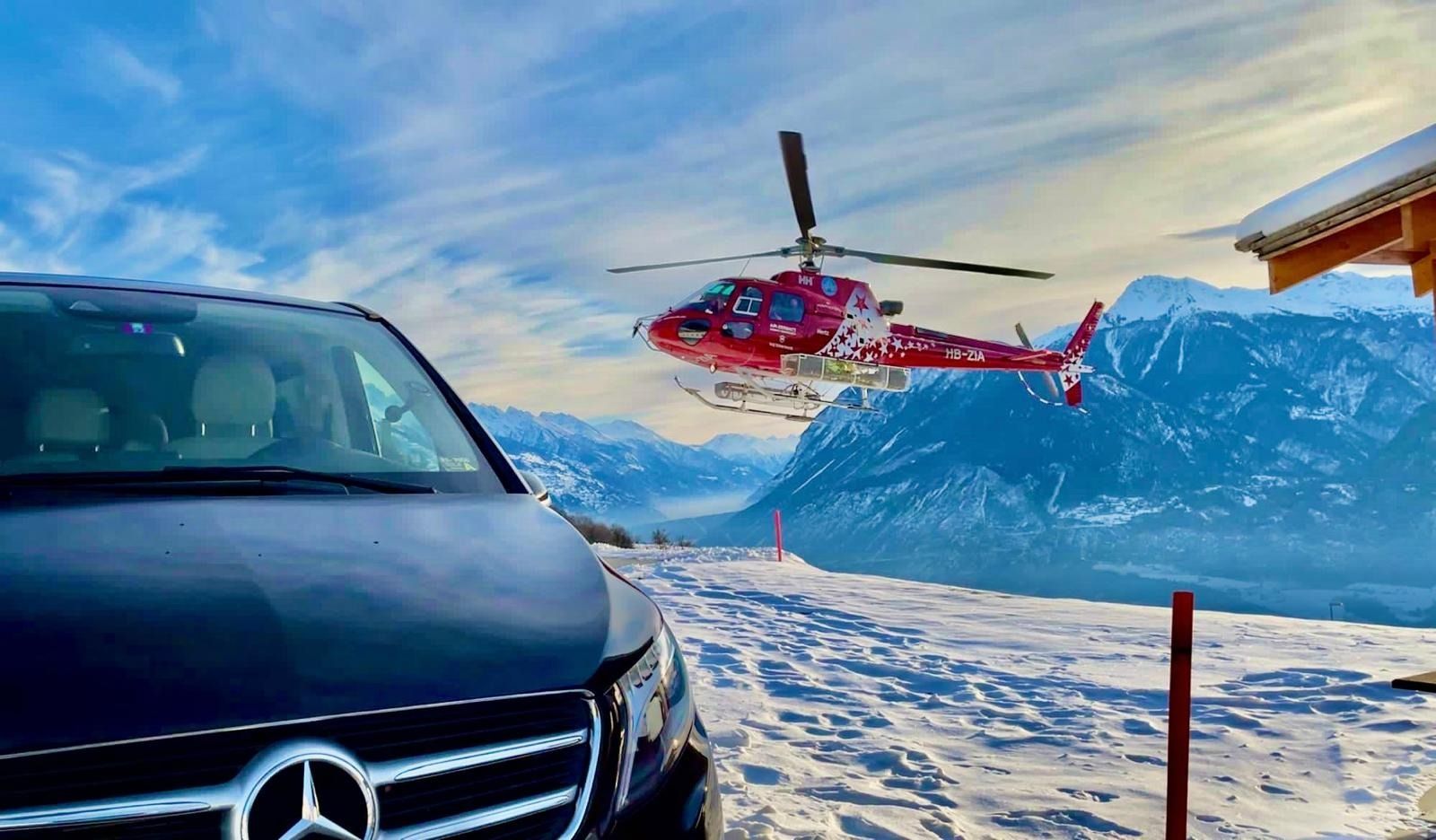 Hélicoptère rouge survolant des montagnes enneigées, camionnette bleue au premier plan.