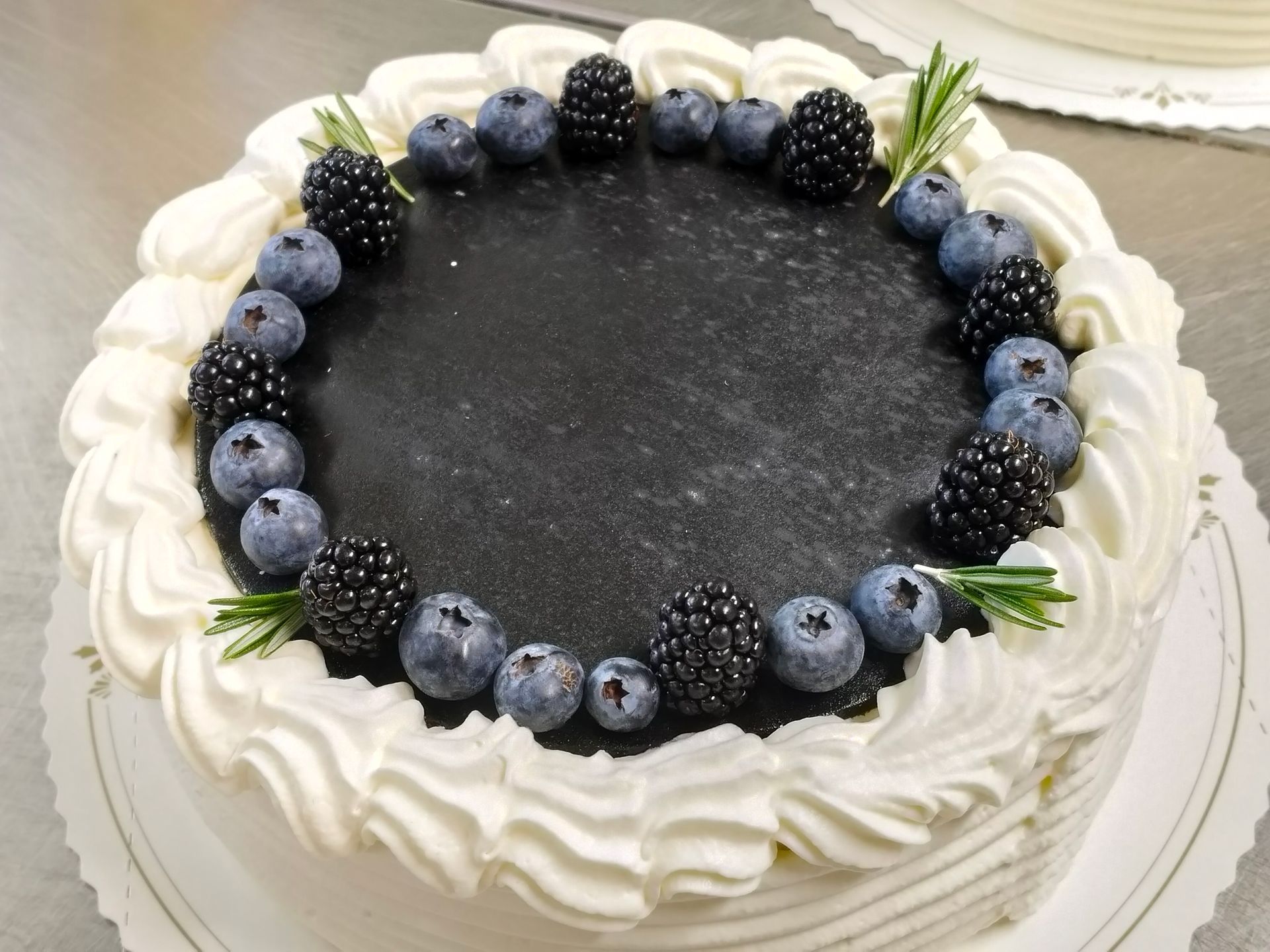 Vit frostad kaka med en mörk mitt och en ring av blåbär, björnbär och rosmarinkvistar