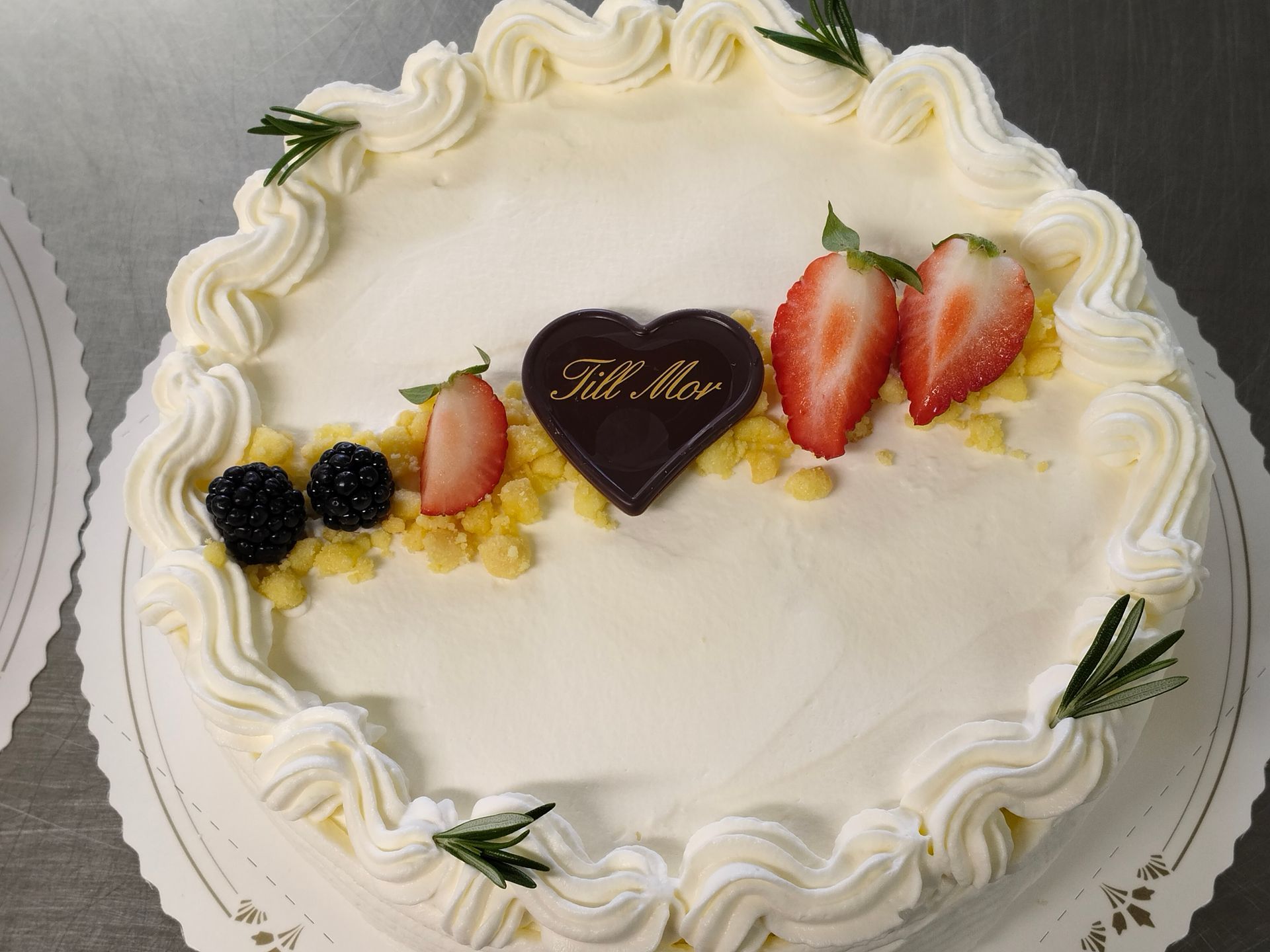 Rund vit kaka med vispad frosting, jordgubbar, björnbär och en chokladhjärta på toppen med texten 
