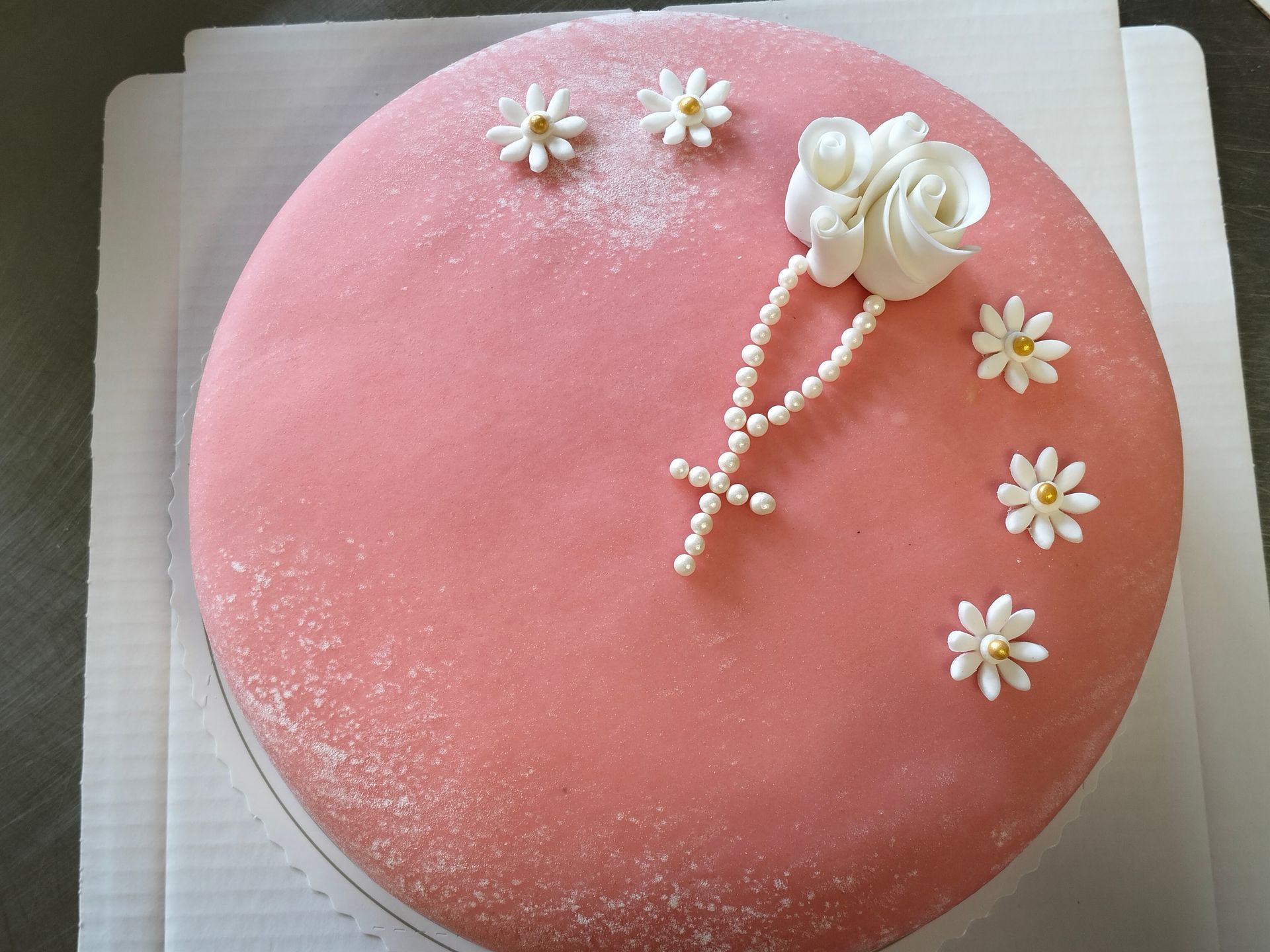 Rosa frostad tårta med vita sockerblommor och en pärlkorsdekoration på toppen