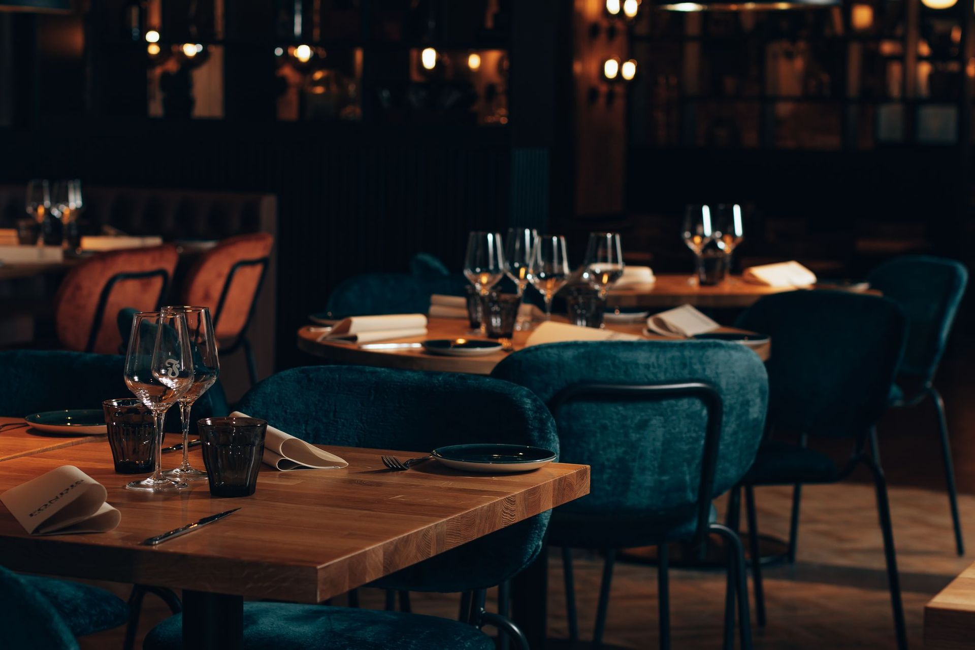 Hämärästi valaistu moderni ravintola, jossa on katetut pöydät, viinilasit ja sinivihreitä tuoleja