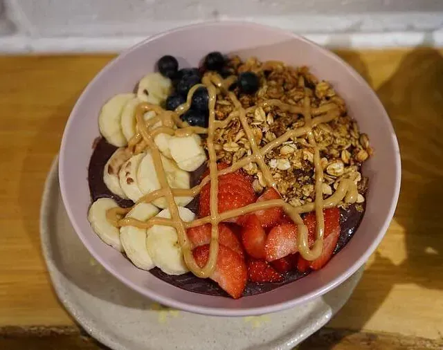 Un tazón de comida con plátanos, fresas, arándanos, granola y mantequilla de maní sobre una mesa.
