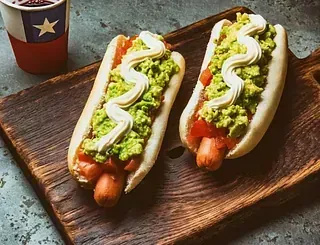 Dos hot dogs con guacamole y mayonesa sobre una tabla de cortar de madera.