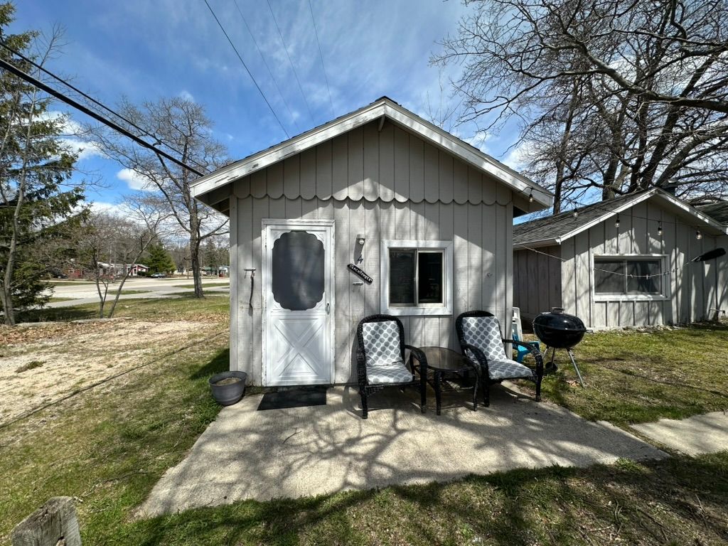 Small beige cottage porch with two chairs, a table, potted plants, and a grill beside a grassy yard.
