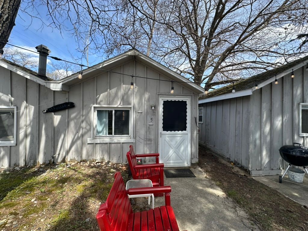 Red Adirondack chairs on a sunlit garden patio beside a small beige cottage with flowers.