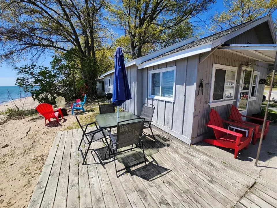 Beachfront cabin deck with red chairs, glass table, and blue umbrella under sunny trees