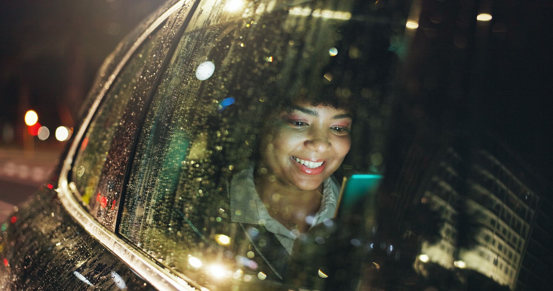 Une personne souriante regarde son téléphone à travers la vitre d'une voiture ruisselante de pluie, la nuit.