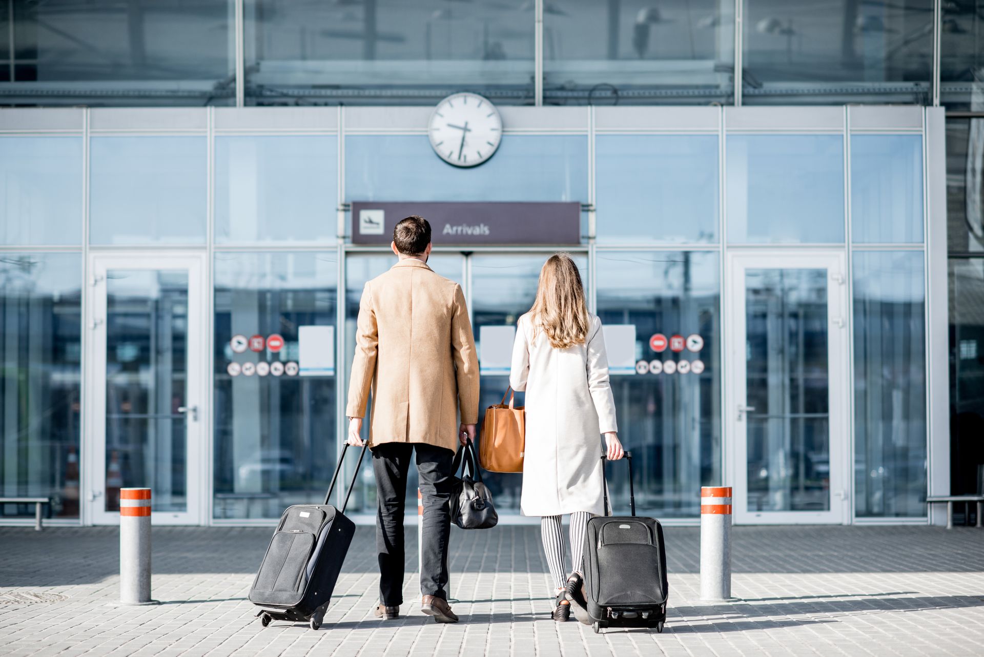Deux voyageurs avec des valises à roulettes se tiennent devant l'entrée d'un aéroport, sous une horloge.