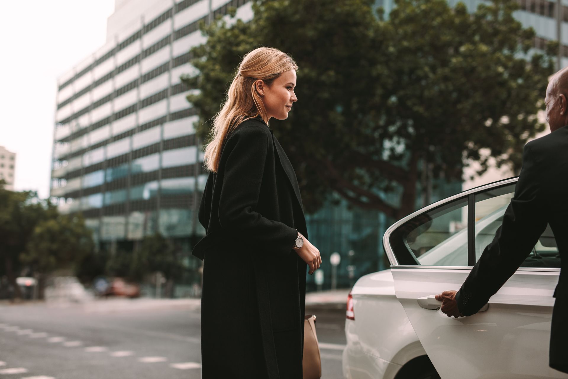 Une femme en manteau noir s'apprête à monter dans la voiture, la portière est tenue par une autre femme.