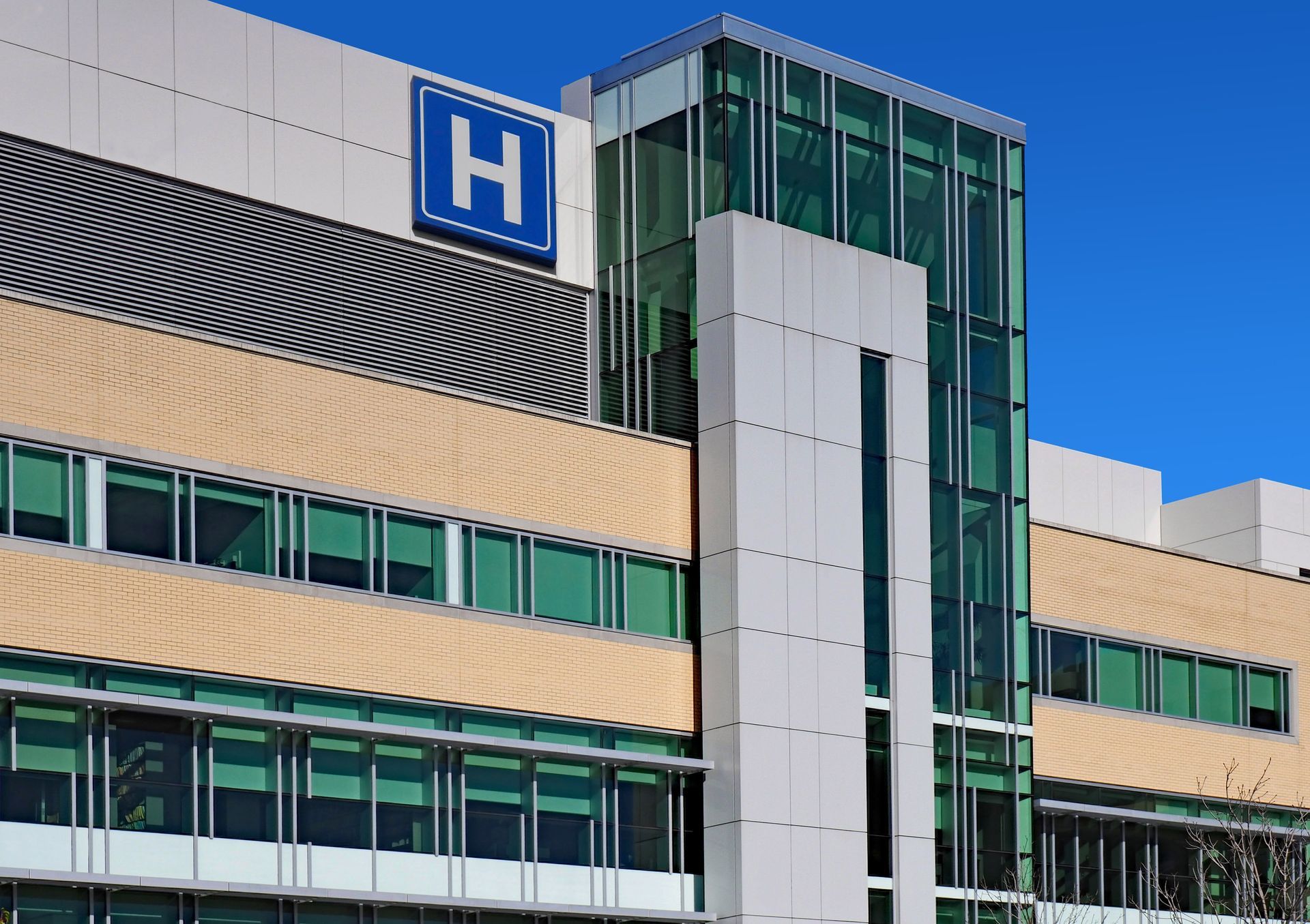 Extérieur du bâtiment de l'hôpital avec le panneau H bleu et la tour d'ascenseur en verre sous un ciel dégagé.