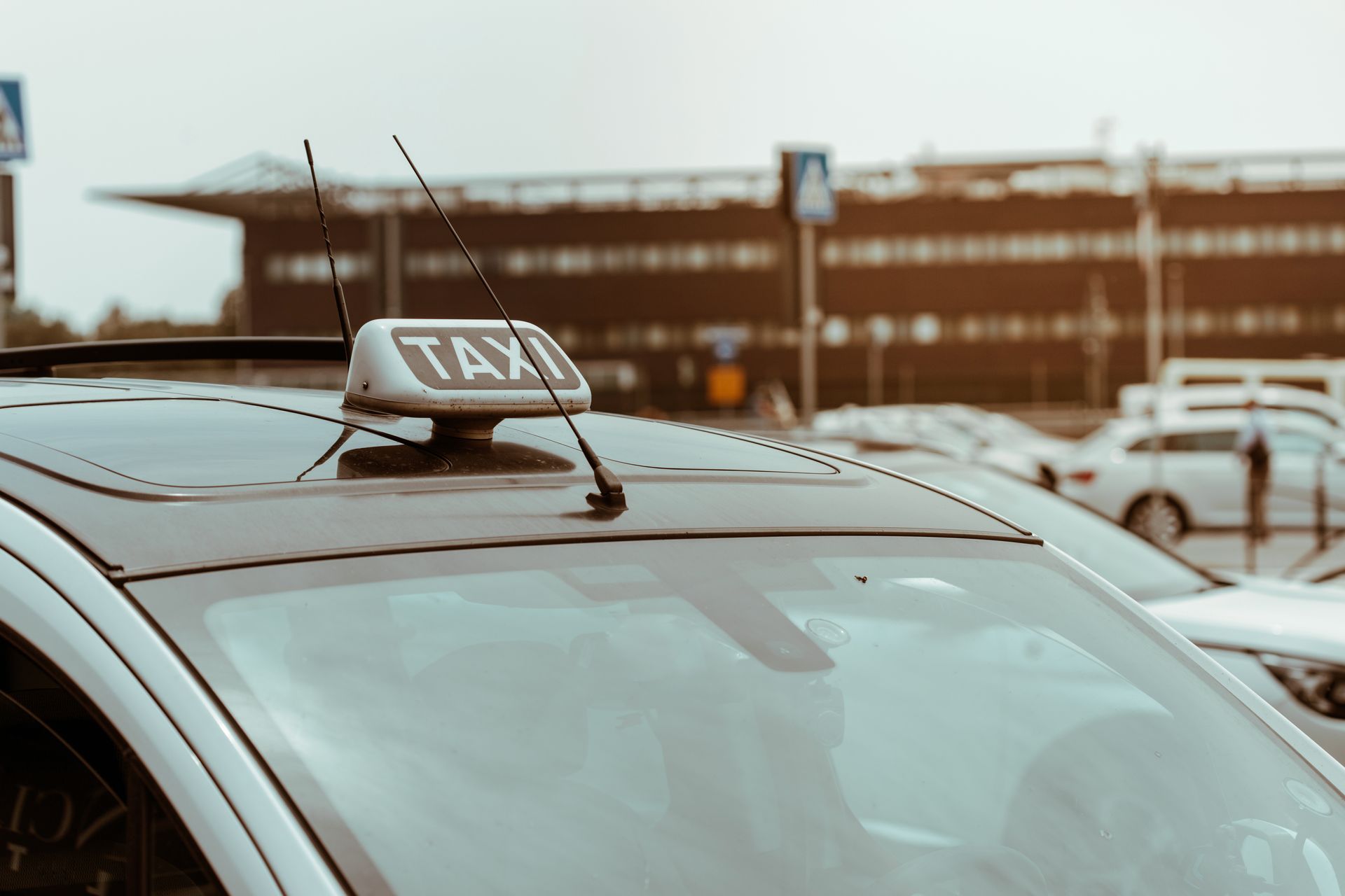 Panneau de taxi sur le toit d'une voiture dans un parking, avec un bâtiment flou en arrière-plan.