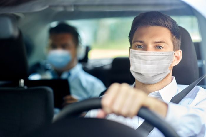 Un conducteur et un passager masqués dans une voiture, l'un concentré sur la route au premier plan.