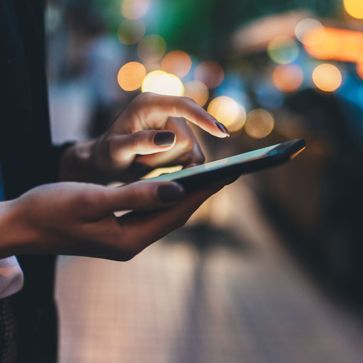 Des mains utilisent un smartphone la nuit, avec des lumières urbaines floues en arrière-plan.