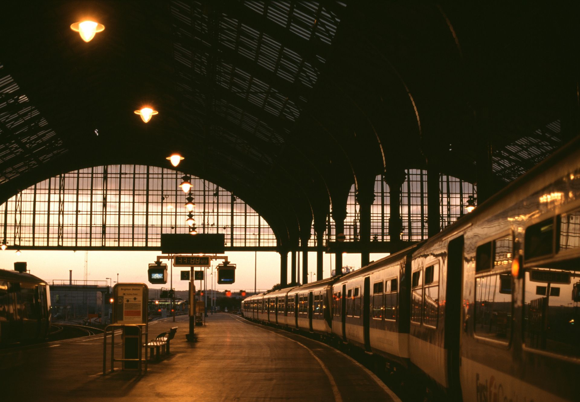 Quai de gare avec une rangée de trains sous une haute verrière en forme d'arche au coucher du soleil.
