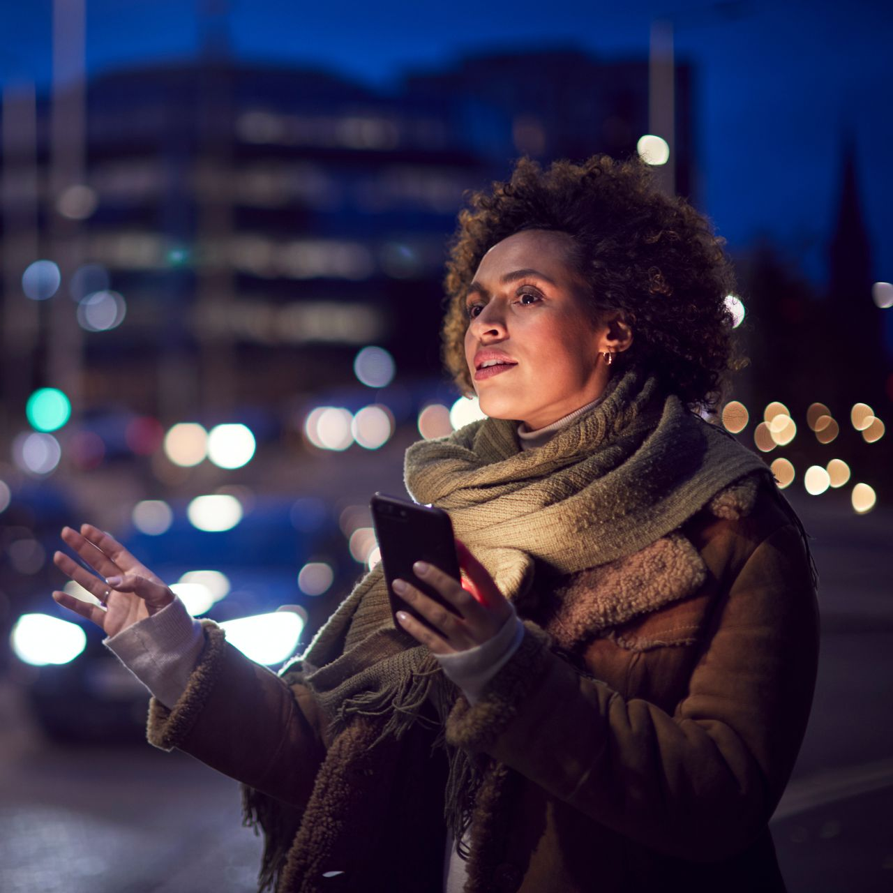 Une personne tenant un smartphone la nuit dans une rue de la ville, avec des feux de circulation et des bâtiments flous en arrière-plan.