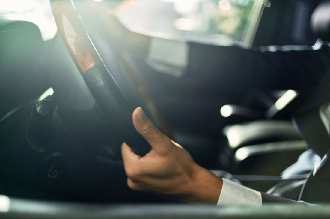 Une main tenant le volant d'une voiture dans un intérieur éclairé par le soleil, avec des reflets vifs à travers le pare-brise.