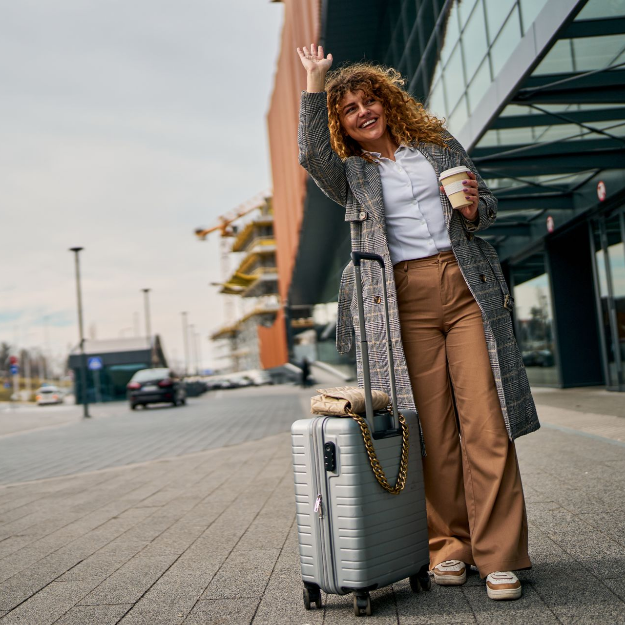 Voyageur souriant et faisant signe de la main à côté d'une valise argentée devant un bâtiment moderne.