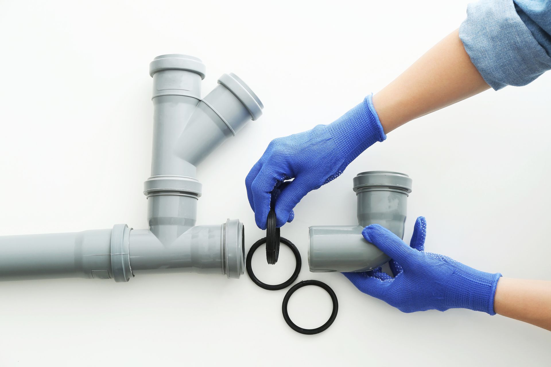 Des mains gantées de bleus assemblent des tuyaux de plomberie en plastique gris et des joints toriques en caoutchouc noir sur une surface blanche.