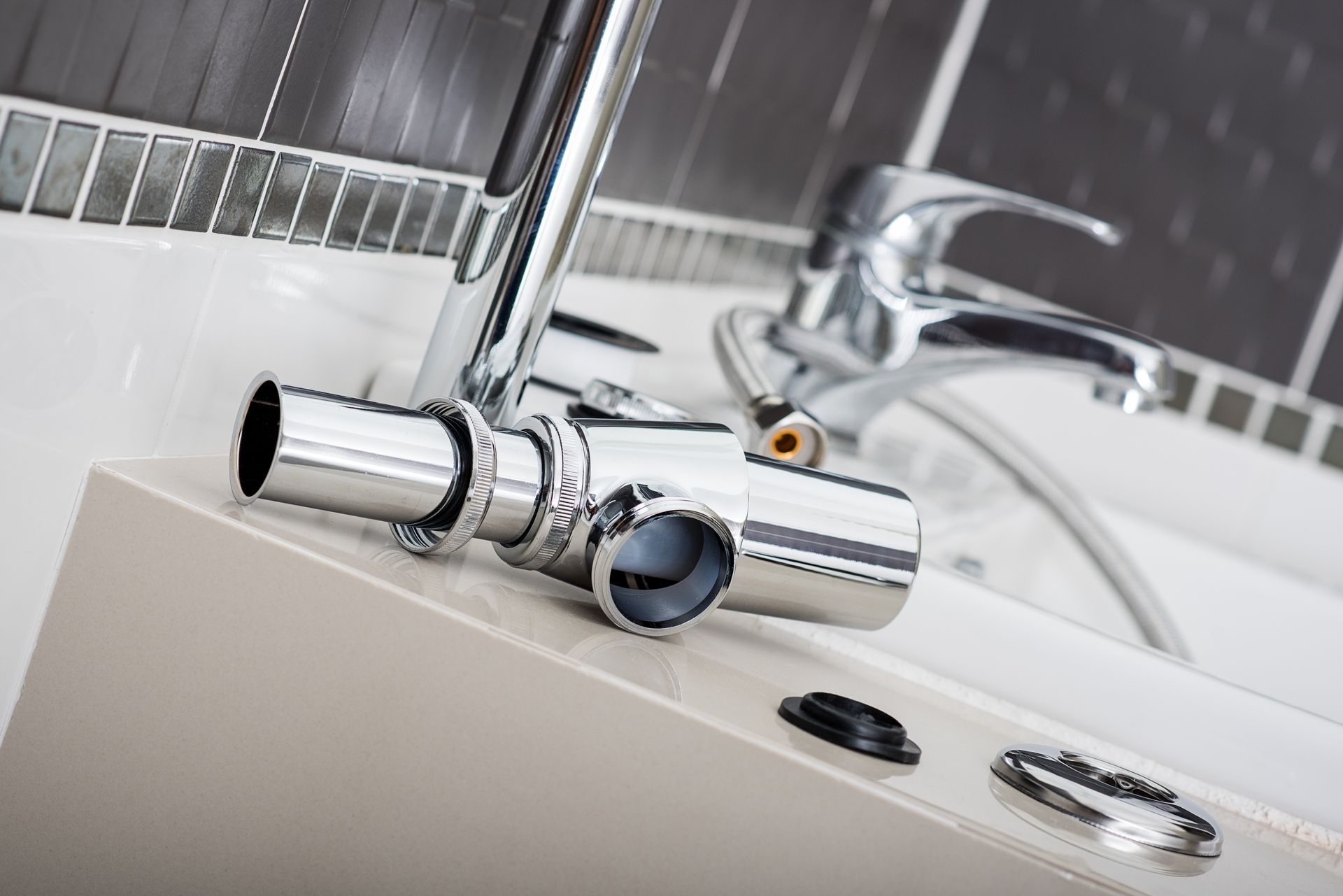 Des éléments de plomberie chromés pour le lavabo et un robinet sont posés sur un plan de travail blanc pour la salle de bains, près d'un mur carrelé.
