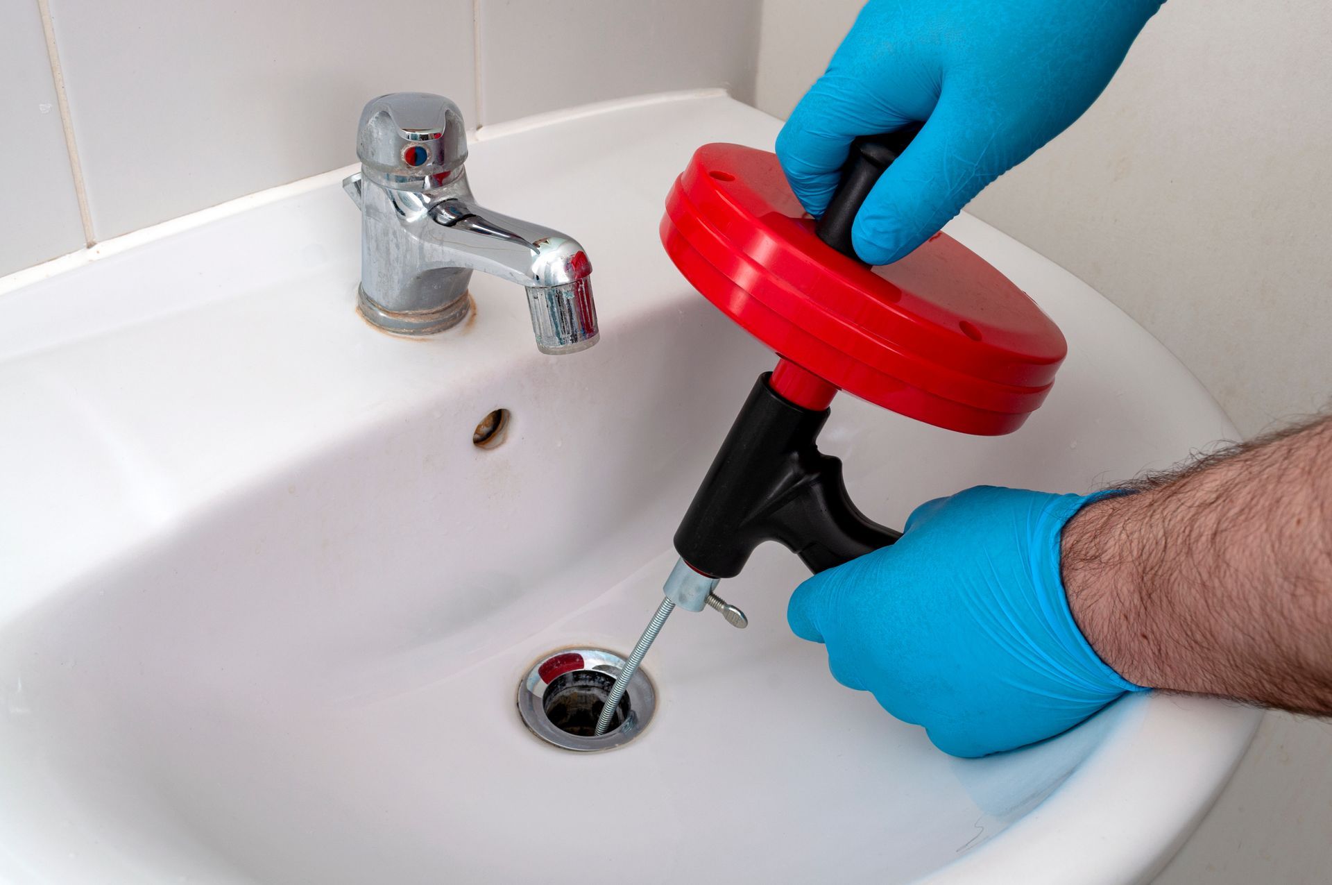 Une personne portant des gants bleus utilise un déboucheur manuel rouge pour déboucher un lavabo blanc.