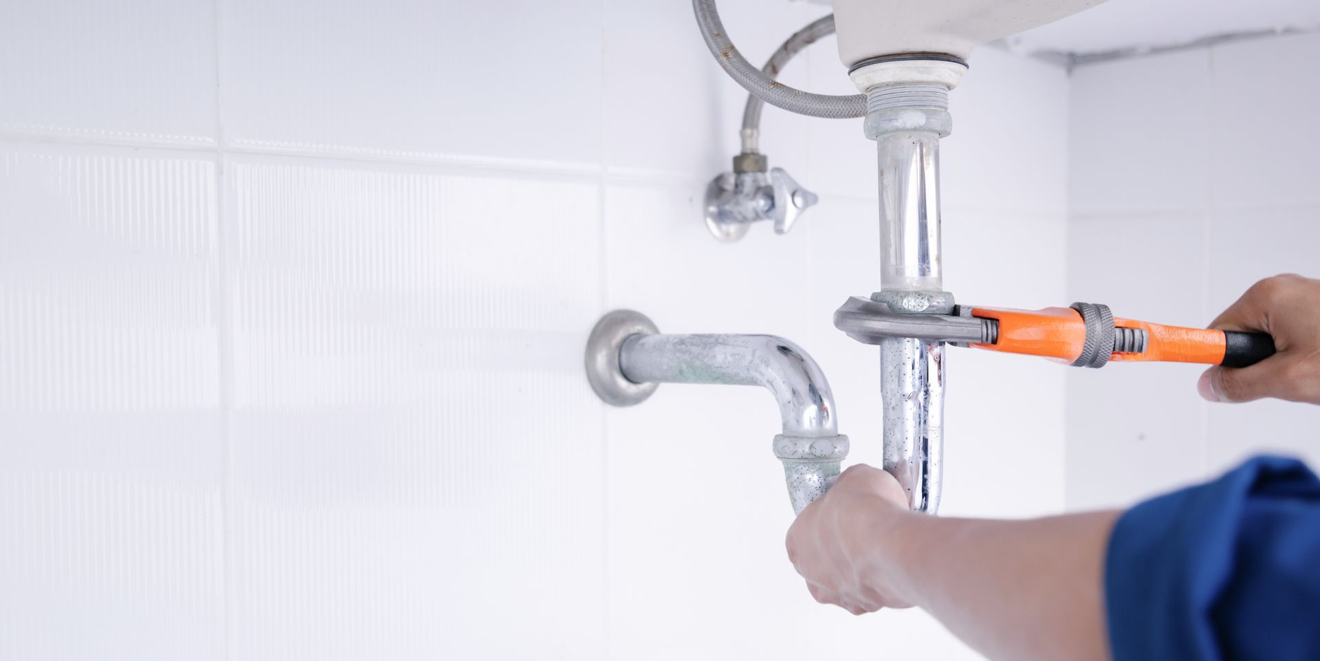 Un plombier utilise une clé orange pour serrer un tuyau métallique sous un lavabo contre un mur carrelé blanc.