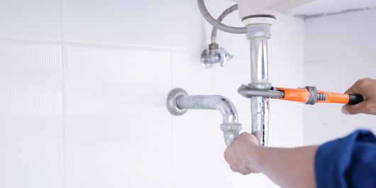 Une personne utilise une clé pour serrer un ensemble de tuyaux métalliques sous un lavabo de salle de bain contre un mur carrelé blanc.