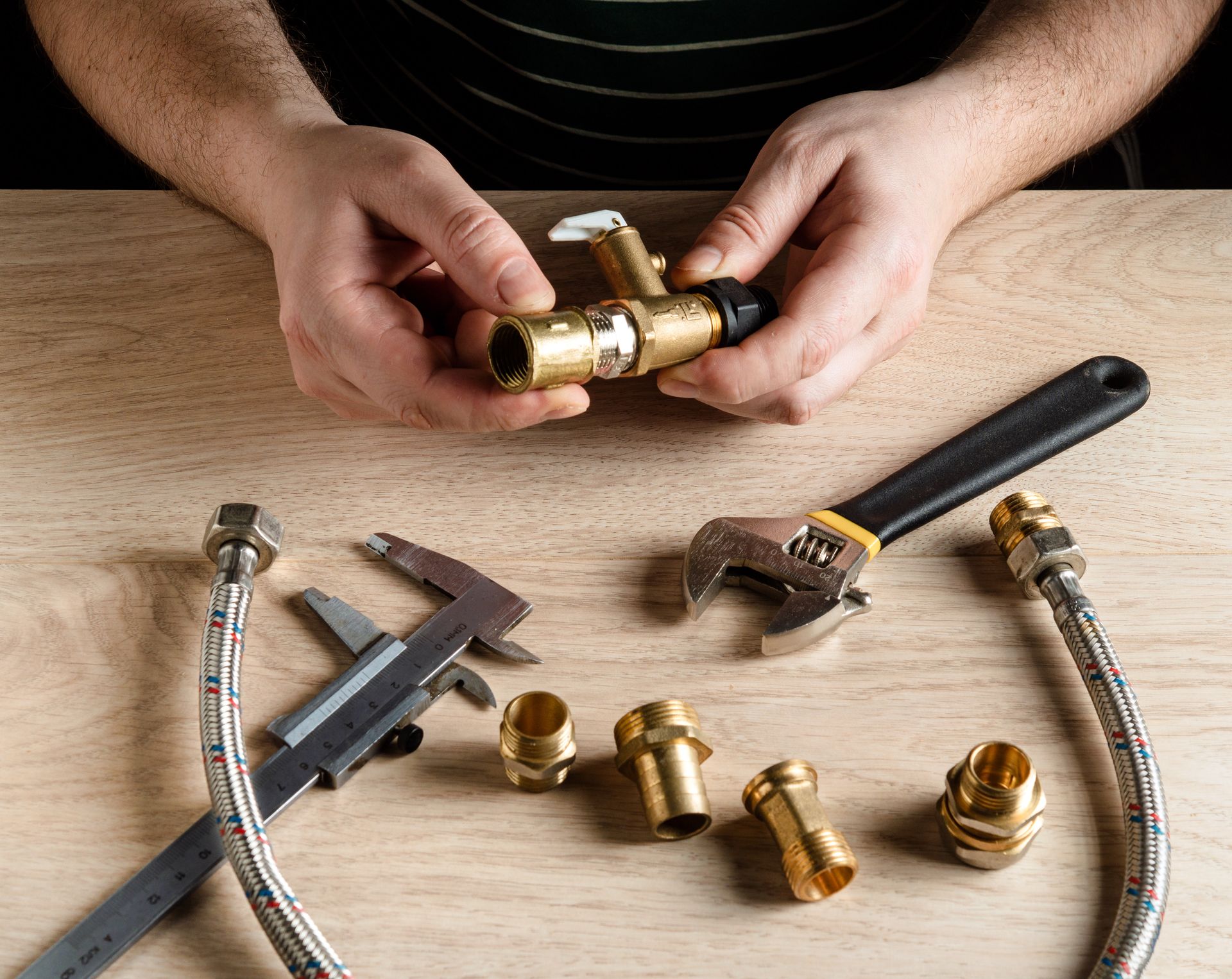 Des mains assemblent des raccords de plomberie en laiton sur une surface en bois à l'aide d'un pied à coulisse, d'une clé réglable et de tuyaux d'alimentation tressés.