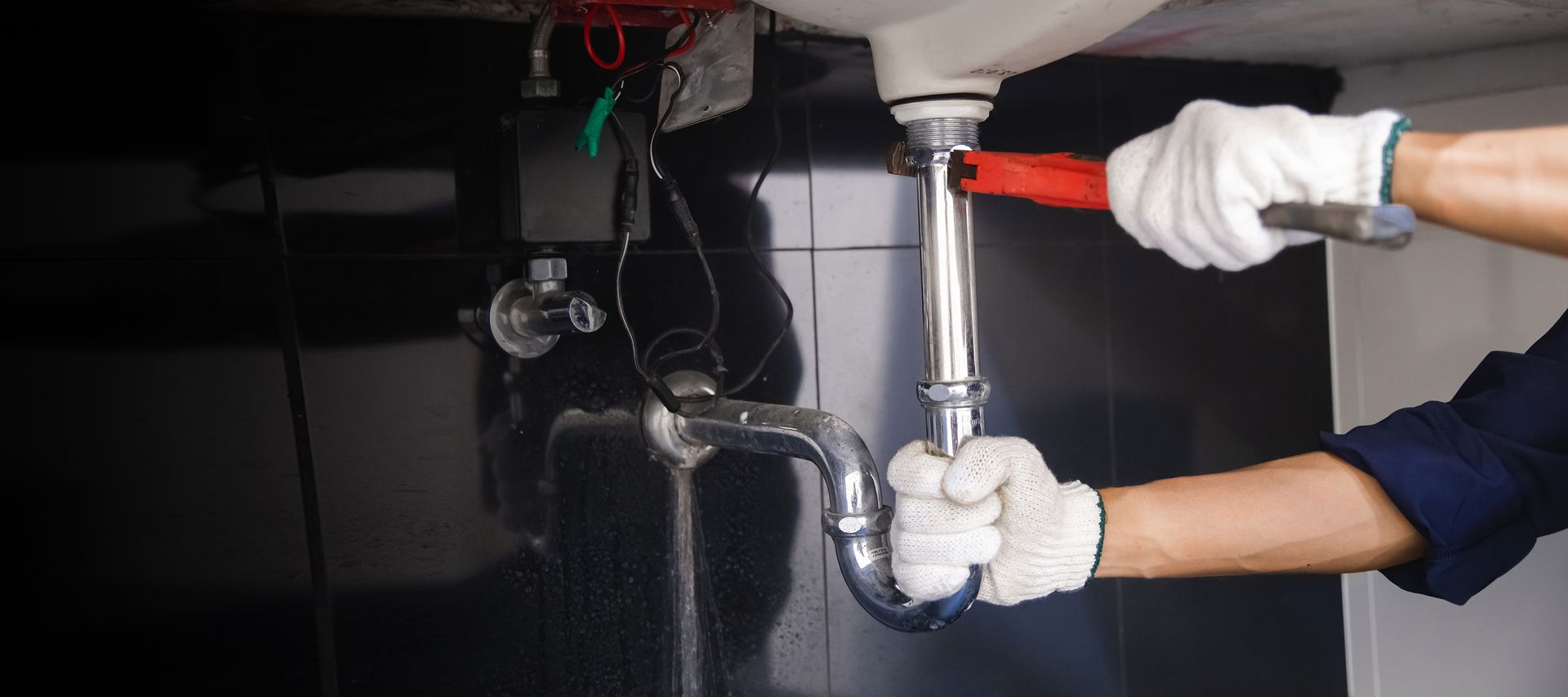 Une personne portant des gants de travail blancs utilise une clé à pipe rouge pour serrer un tuyau d'évacuation chromé sous un évier.